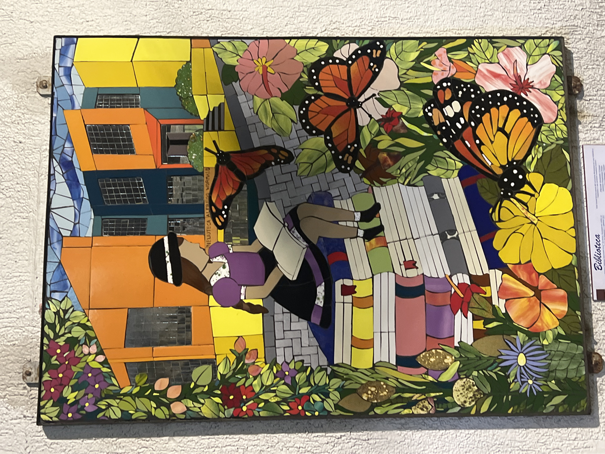 A mosaic mural attached to a white textured wall. The artwork depicts a young woman with dark hair, a headband, a purple shirt, and a black skirt. She is reclining on a stack of books and holding another open book. The mural has the artistic style of mosaic tiles and includes architectural elements: a building with yellow, orange, and blue facades. There are also representations of flowers and monarch butterflies. The word "Biblioteca" is written on a small sign next to the mosaic. It's likely located in Arica, Chile given the prompt.