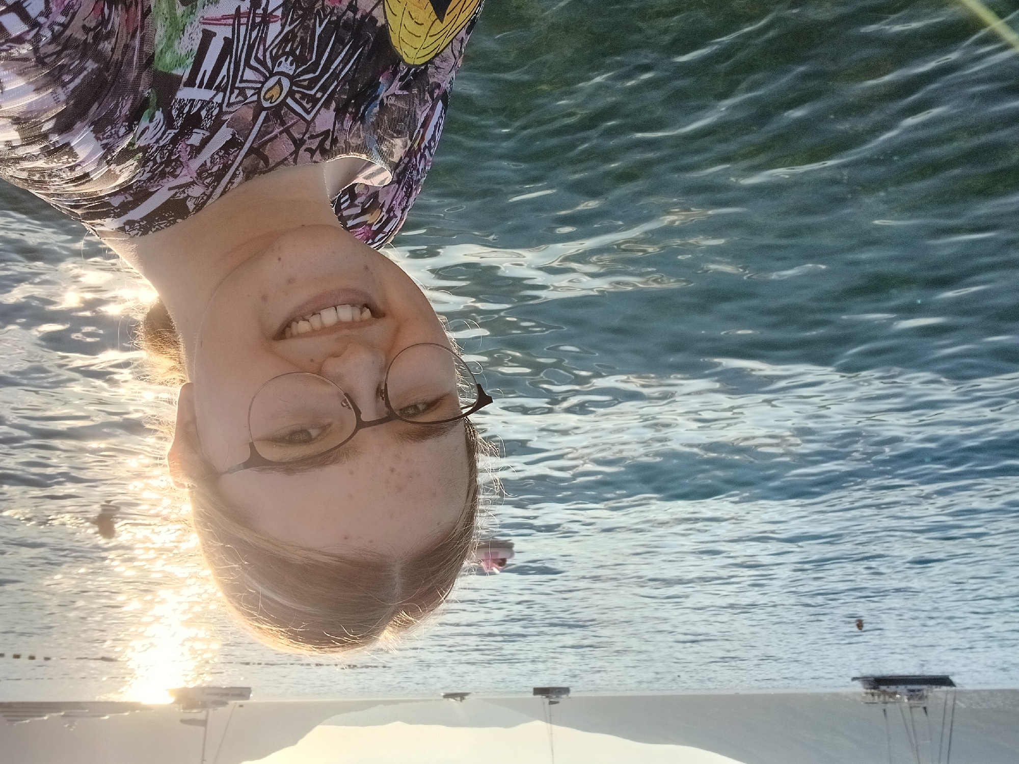 A young woman with blonde hair and glasses is taking a selfie outdoors on a body of water. She is smiling at the camera and wearing a colorful shirt. In the background, there are boats and a hazy mountain range. The sun is shining brightly, reflecting off the water. The environment appears to be a coastal or lake area.