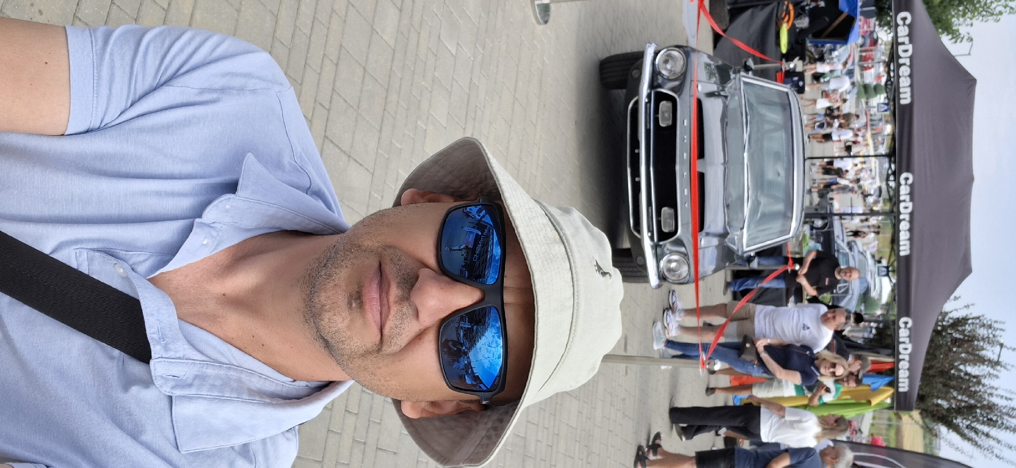 A man wearing a light blue polo shirt, a light-colored bucket hat, and dark sunglasses is taking a selfie outdoors. He is standing in front of a classic black car, likely a Shelby Mustang, which is cordoned off with red tape. Behind the car, tents with "CarDream" branding are visible, suggesting an outdoor event like a car show or exhibition. Several other people are present in the background, some looking at the car or mingling. The ground is paved with light-colored square stones. The overall scene suggests a public gathering in Stăuceni, Moldova, focused on automobiles.