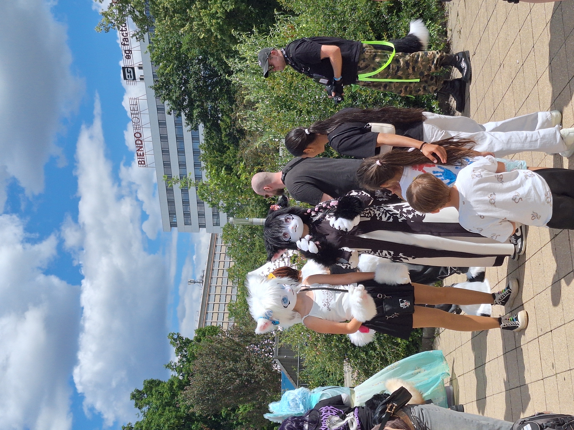 The image captures an outdoor gathering on a paved area, likely a public space in Chemnitz, Germany, with a building labeled "BIENDO HOTEL" visible in the background. Several individuals are present, including some dressed in elaborate furry costumes, featuring animal-like headwear and full-body suits. One person wears a white cat-like costume with large blue eyes and fluffy white accessories. Another is in a black and white striped outfit with a furry black hood and paws. A young boy in a white t-shirt stands facing away from the camera, while a girl with a ponytail stands beside him, holding onto his arm. Several adults are also visible, some observing the costumed individuals. A man in camouflage pants and a black shirt, holding a camera, is positioned towards the right side of the frame. The environment is characterized by green trees and shrubs, with a bright blue sky dotted with white clouds overhead. The overall scene suggests a convention, festival, or organized fan event.