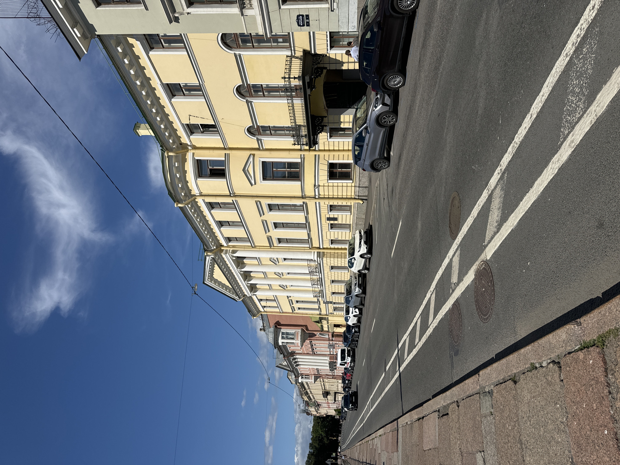 The image shows a street scene in Saint Petersburg, Russia. The street is lined with multi-story buildings, most of which are painted in light colors, with classical architectural features such as columns and arched windows. Cars are parked along the side of the road and driving on it. The sky is blue with some scattered clouds. The sidewalk is made of stone. There is a street sign visible on one of the buildings with Cyrillic text. The overall impression is of a sunny day in a European city.