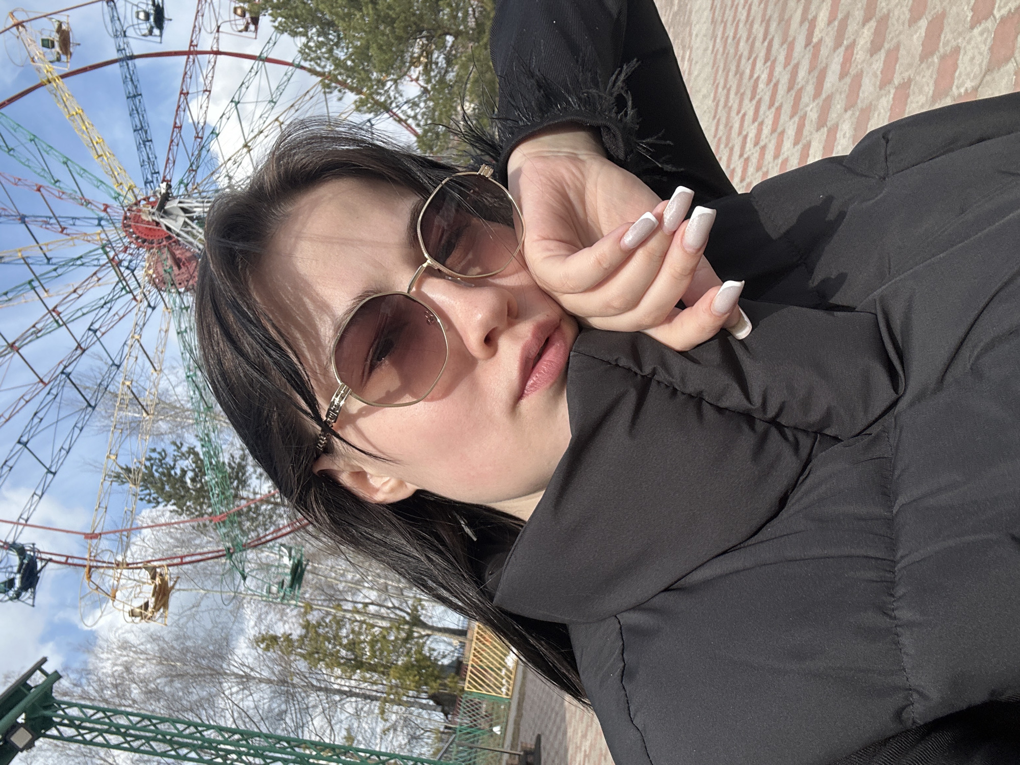 A medium close-up, eye-level shot features a young woman posing for a selfie in an outdoor setting, likely an amusement park in Chaikovsky, Russia. The woman, with dark hair, wears large, gold-framed, hexagonal-shaped sunglasses and a black puffy jacket. Her right hand, adorned with long, square-tipped nails featuring a white-tipped design, rests gently against her face or jacket collar, drawing attention to her manicured fingers. Her lips are slightly parted, revealing a natural pink hue. The black jacket has a visible seam line and appears to have a textured or feathery cuff on the sleeve near her hand.
In the background, a large, colorful Ferris wheel dominates the upper left portion of the image, showcasing its intricate metal structure with sections painted in red, yellow, and blue against a bright blue sky with scattered white clouds. The bare branches of trees suggest early spring or late autumn. Other fragmented structures, possibly from additional amusement park rides, are visible around the Ferris wheel. The ground to the lower right shows a patterned paved surface, possibly made of light-colored bricks or tiles. The overall lighting indicates a bright, sunny daytime setting. There is no visible text in the image.