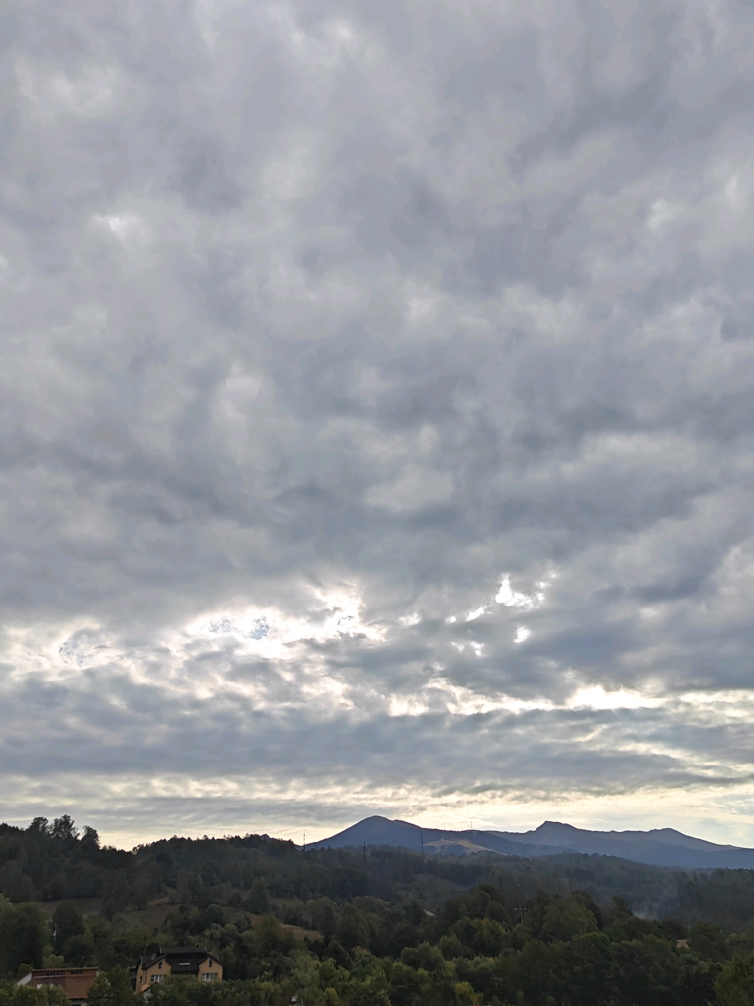 The image depicts a landscape scene under a cloudy sky. The foreground shows a dense forest with several houses scattered among the trees. The houses appear to be of traditional design, with pitched roofs. In the middle ground, rolling hills covered in trees are visible, with some structures, possibly communication towers, dotting the landscape. The background features a mountain range under a dramatic sky filled with various cloud formations. Patches of sunlight break through the clouds, illuminating parts of the scene. The overall impression is one of a rural or semi-rural setting with natural beauty.