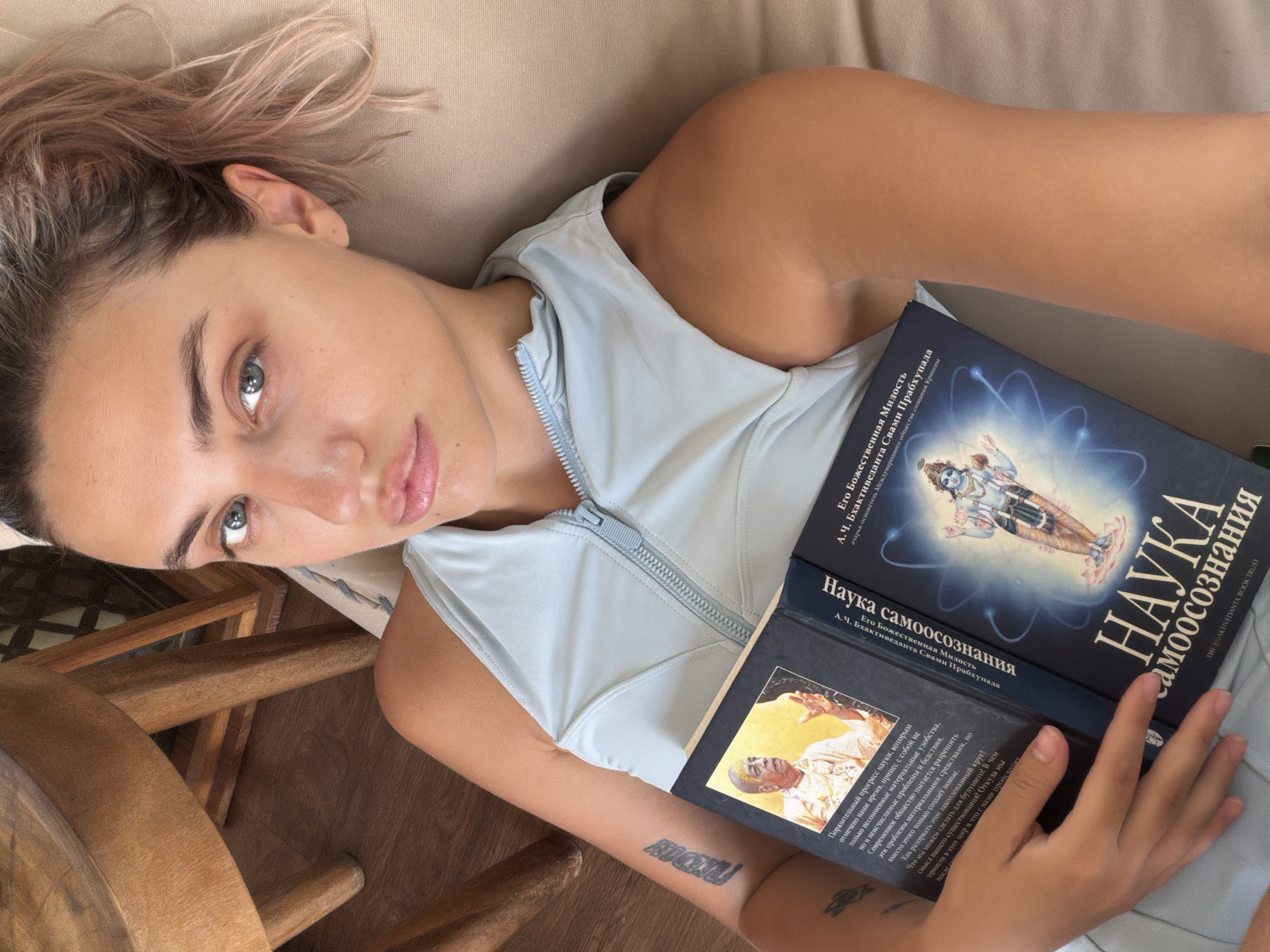 A young woman with blonde hair pulled back in a ponytail and light pink streaks lies on her side, holding an open book in her hands. She is wearing a light blue, zippered tank top. The book's cover features an illustration of a Hindu deity and text in Russian. The woman's left arm has a tattoo that reads "freedom" and another smaller tattoo below it. She is looking directly at the camera with a neutral expression. The background appears to be a dimly lit room with wooden furniture, suggesting an indoor setting. The image appears to be a selfie taken from a high angle. The location is noted as North Kuta, Indonesia.