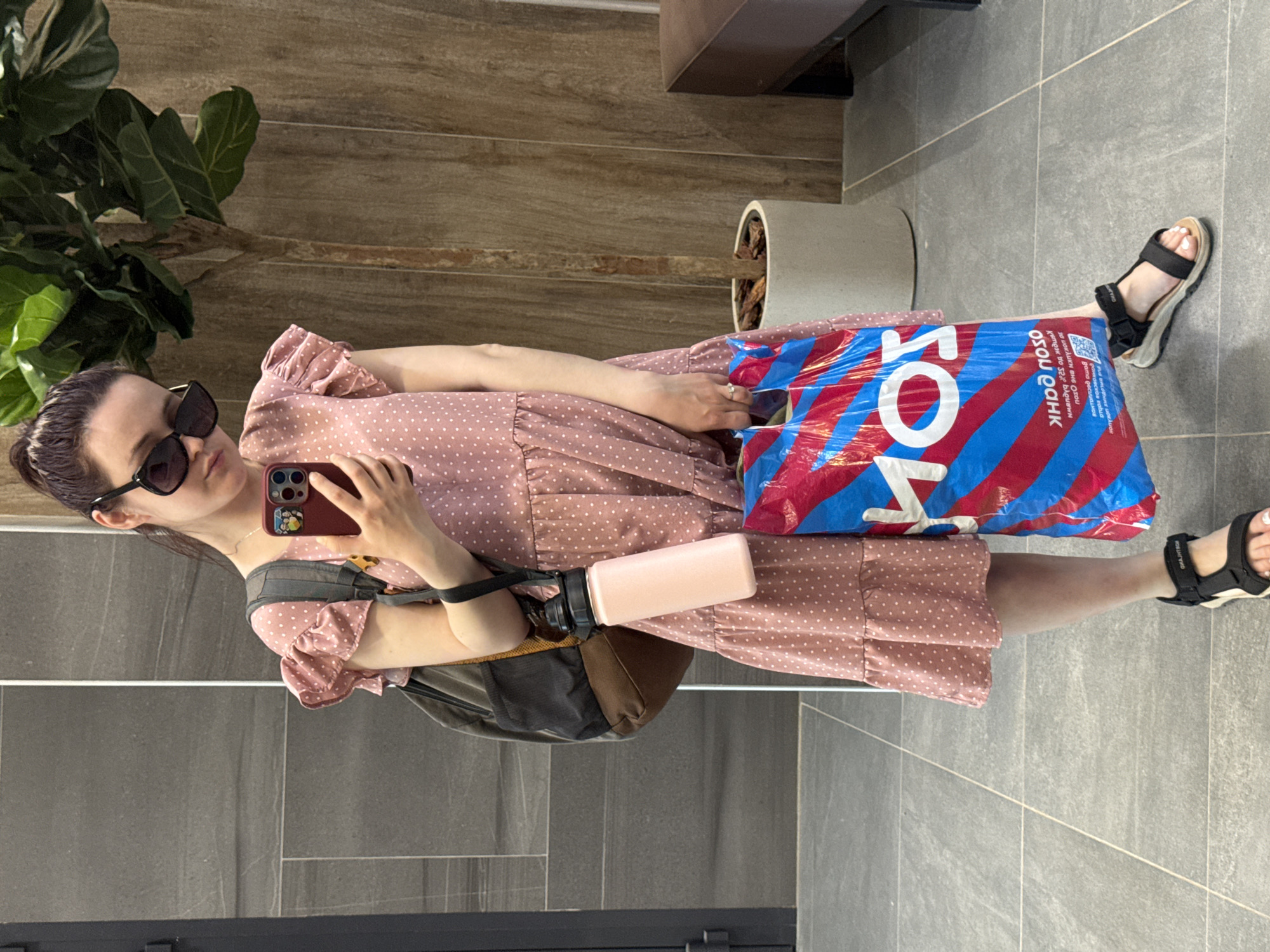 A young woman stands indoors, holding a smartphone and taking a selfie. She is wearing a pink polka-dot dress and black sandals, with a backpack slung over her shoulder. In her other hand, she carries a large blue and red plastic bag with Cyrillic text on it, indicating she has recently been shopping. A pink water bottle is tucked into the straps of the backpack. The background includes tile flooring, a wooden wall panel, and a potted plant, suggesting a public space, possibly a shopping mall or apartment building entrance. Based on the Cyrillic text and the presence of a bag from a prominent Russian retailer, the location is likely Izhevsk, Russia.