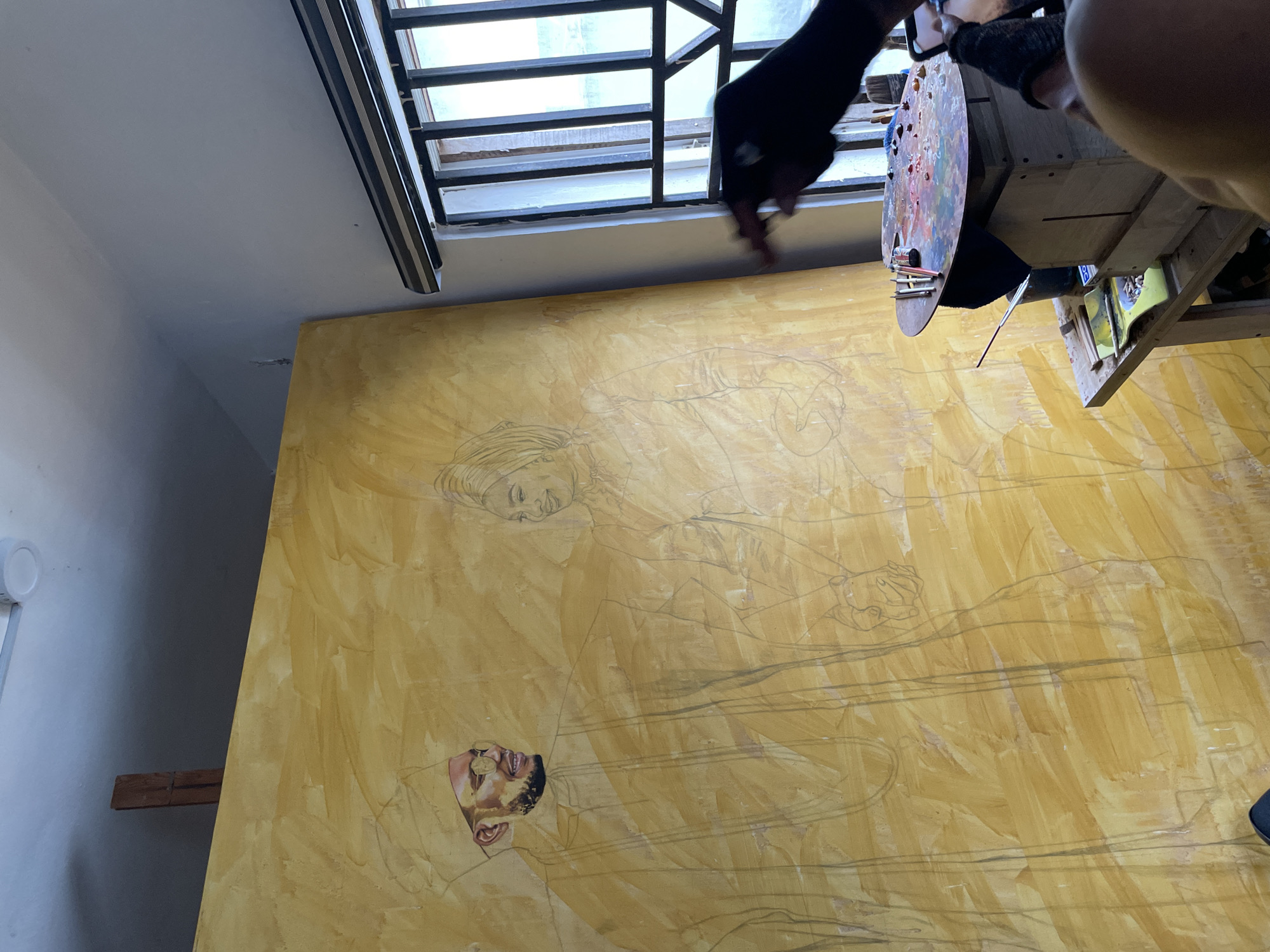 The image shows an artist painting a large canvas in a room with a window near the ceiling. The canvas is predominantly yellow with sketches of two figures, one near the top of the canvas and the other near the bottom. The artist is holding a paint palette and brush. The location is Ado, Nigeria.