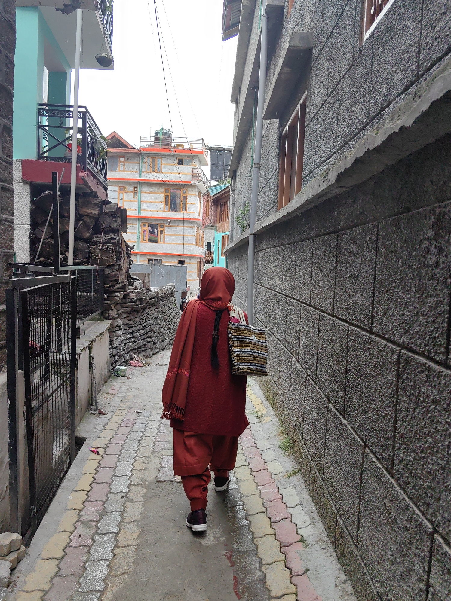 A person wearing a red headscarf and matching red salwar kameez walks down a narrow alleyway paved with multicolored interlocking blocks. They carry a striped woven bag over their shoulder. To the right, a rough, dark gray stone wall forms the side of a building. To the left, a metal fence encloses an area with stacked firewood and a turquoise building with a balcony. In the background, a multi-story building with orange and white siding is visible. The scene appears to be in the city of Vashisht, India.