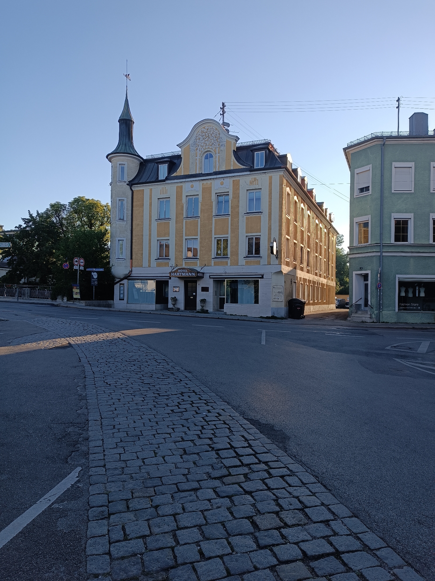 The image shows a street scene in Fürstenfeldbruck, Germany, featuring a yellow and white building with the sign "HOTEL HARTMANN HOTEL" and a tower. To the right, a light green building with white trim and window shutters is visible, displaying a sign for "Fashion Store". The street is paved with cobblestones in the foreground, transitioning to asphalt. The sky is clear and blue, suggesting a daytime setting with the sun casting shadows. There are street signs and traffic signs present, including a parking sign. Trees are visible in the background to the left of the yellow building. The scene appears to be an urban environment.