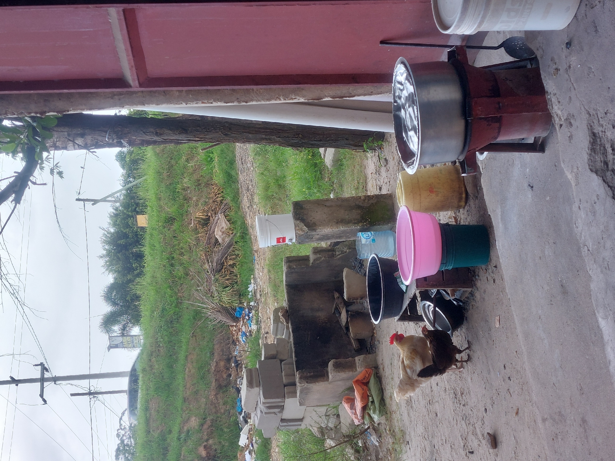The image shows an outdoor cooking setup. A large metal pot sits on a red, homemade stove. There are several plastic buckets and containers nearby, including a pink one. A charcoal-burning outdoor stove made of bricks is also visible, with some trash scattered around it. Two chickens are present in the scene. In the background, there is a grassy hill, a telephone pole, and some trees. The scene appears to be located in a semi-rural or suburban area. The specific location within Dar es-Salaam, Tanzania, is not determinable from the provided image.