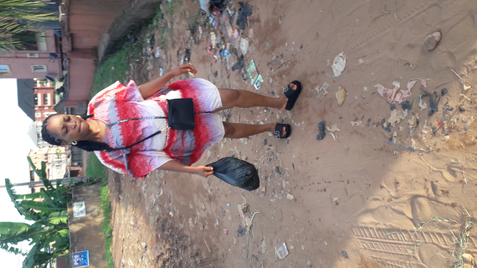 A medium, eye-level shot depicts an adult woman in Nkpor, Nigeria, standing on a dirt road littered with trash. The woman, dark-skinned with braided hair, wears a red and white dress, black sandals, and a black crossbody bag. She holds a black plastic bag in her left hand. The road is mostly dirt with scattered refuse, including paper, plastic, and other debris. In the background, a building and vegetation, including banana trees, are visible. The area appears to be a less developed part of the city. A sign on the bottom left is partially obscured, with the letters "POS" visible. Tire tracks are noticeable in the dirt road.