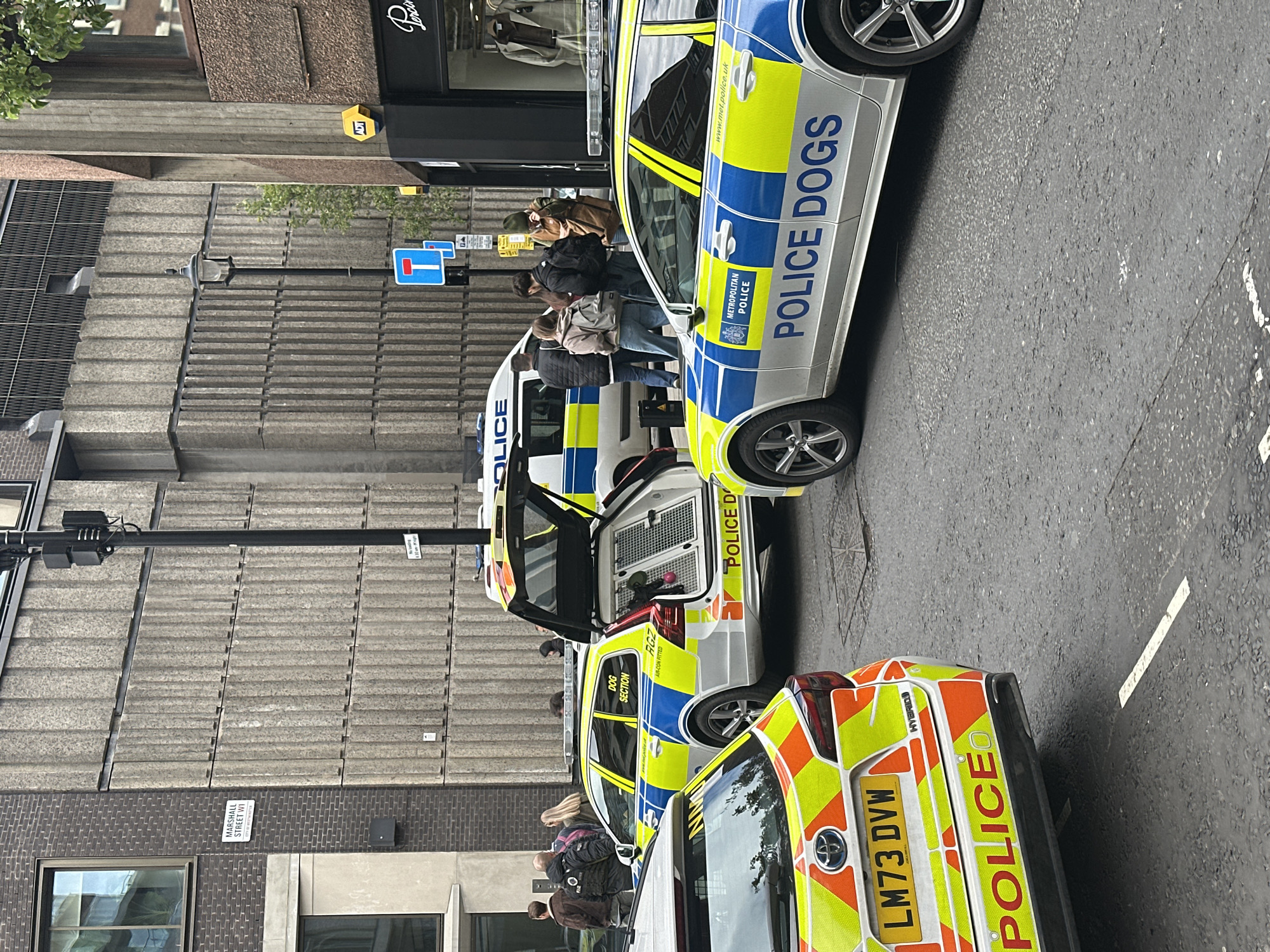 The image shows a street scene in the city of London, United Kingdom. Several Metropolitan Police vehicles, marked "POLICE DOGS," are parked on the street. One vehicle's rear door is open, revealing the interior. Pedestrians are present near the police vehicles. Street signs indicate "MARSHALL STREET WI." The registration number of the first Police vehicle from the bottom of the picture is LM73 DVW.