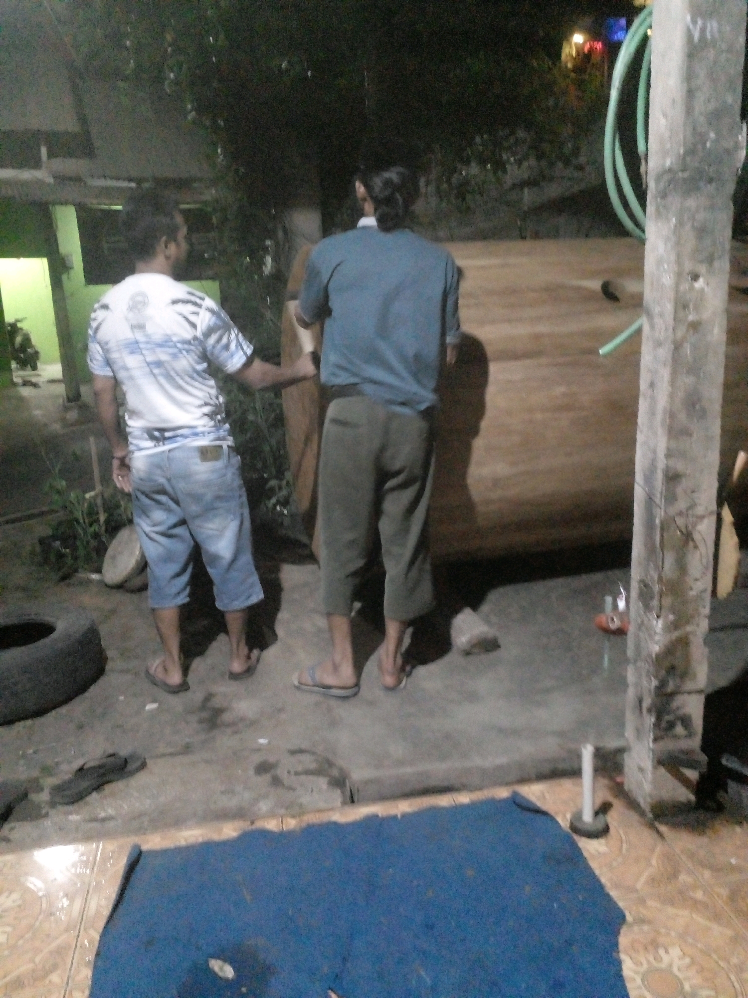 In the city of Pungangan, Indonesia, two individuals are observed near a large, hollow wooden cylinder. The person on the left, wearing a white and blue striped t-shirt and denim shorts, stands with their back to the camera. The individual on the right, in a blue t-shirt and gray pants, is facing away and partially obscured by the cylinder. A green hose is visible in the background, coiled near a concrete pillar. The ground is a mix of concrete and tiled surfaces. A dark blue rug lies in the foreground, partially covering the tiled floor. A discarded tire is also present on the left side of the image. The scene appears to be outdoors, possibly in a workshop or residential area, with trees and a building visible in the dimly lit background.