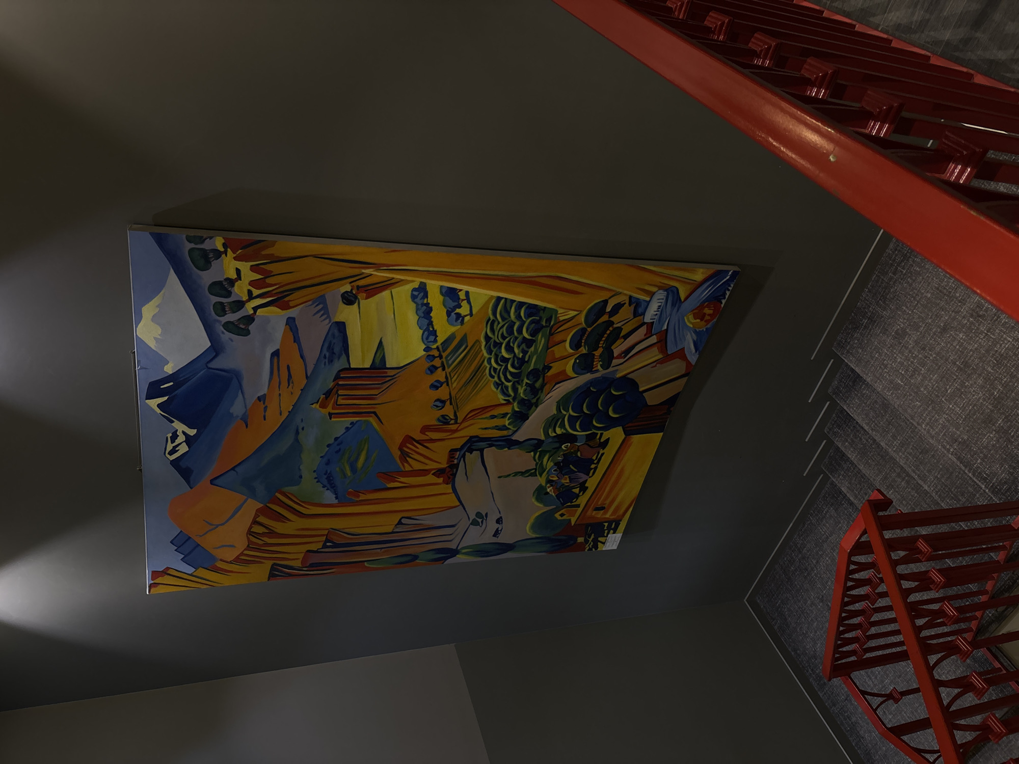 A painting hangs on a gray wall next to a red staircase in Yerevan, Armenia. The painting depicts a colorful, abstract landscape of mountains and trees. The staircase has gray carpet and red railings.
