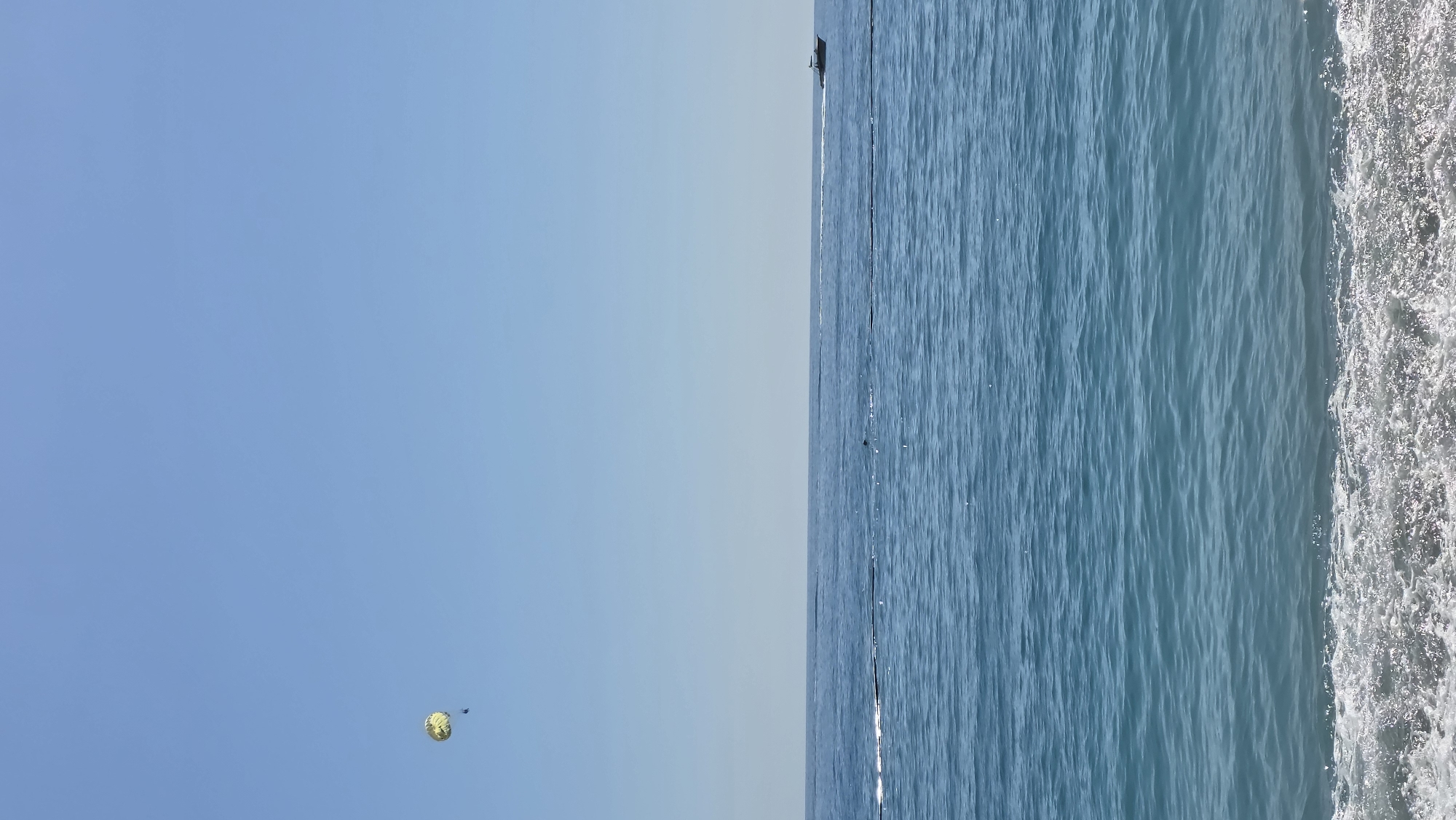 The image, correctly oriented, presents a clear blue sky occupying the upper half of the frame, meeting the calm, deep blue sea that fills the lower half. In the upper left portion of the sky, a bright yellow and orange parasail is distinctly visible, with one or two small dark figures (people) suspended beneath it. On the horizon line, where the sky meets the sea, a small, dark motorboat is seen moving across the water, likely towing the parasail.
The sea's surface is textured with gentle ripples and reflects the light, appearing slightly darker towards the horizon and lighter closer to the foreground. Along the far right edge of the frame, a band of white, frothy water indicates either small waves breaking or the wake of a boat passing nearby. The scene depicts a bright, sunny daytime setting, suggesting excellent weather conditions for water sports and recreational activities. No text is visible in the image. The elements are characteristic of a leisure activity in a coastal region like Fethiye, Turkey.