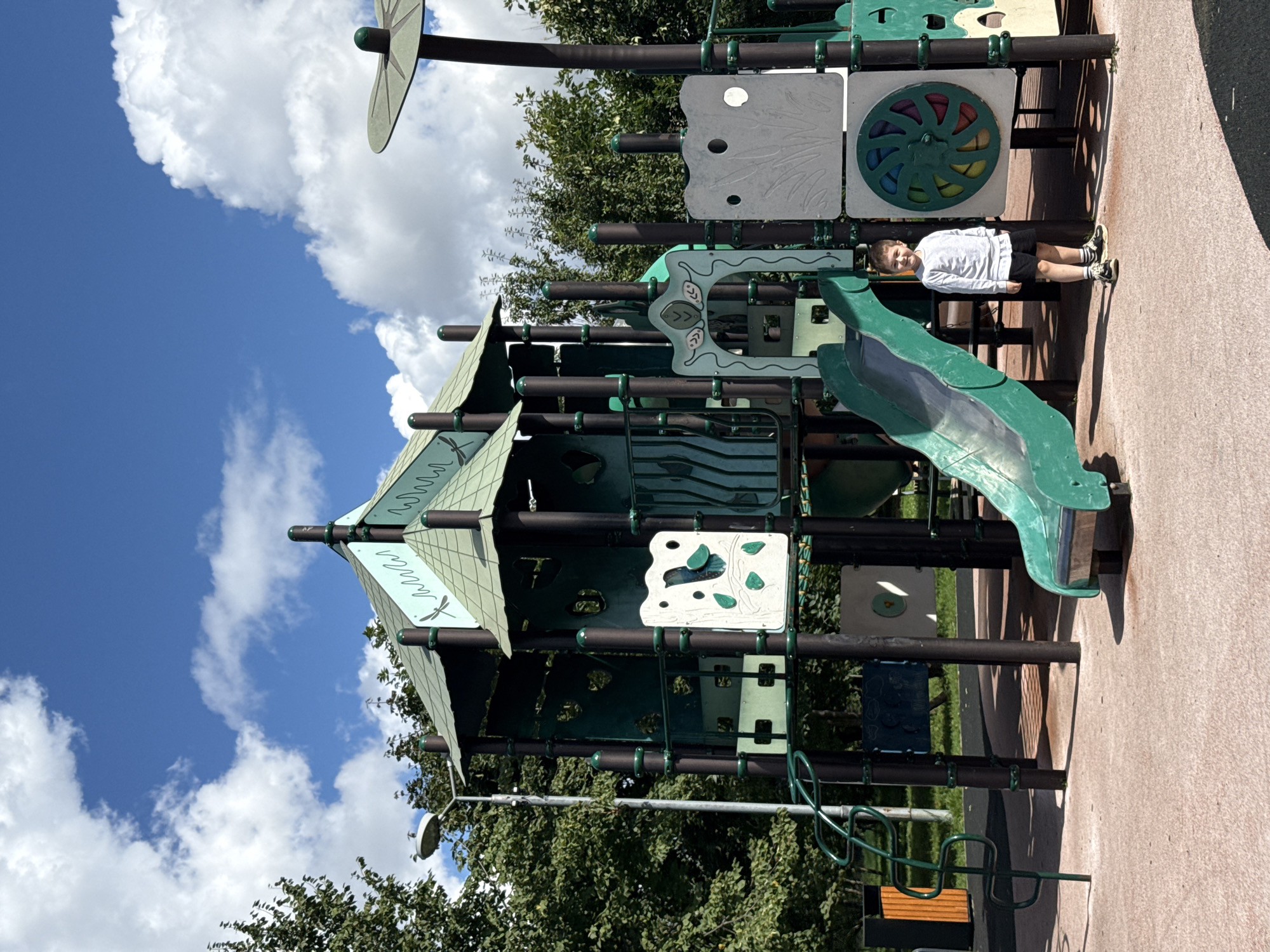 A young boy, estimated to be between 5-7 years old, stands on the rubberized playground surface in front of a large, multi-level green and teal playground structure. He is wearing a light gray long-sleeved shirt and black shorts with striped socks and dark shoes. The playground features multiple slides, climbing elements, and decorative panels with nature motifs. The sky above is blue with scattered white clouds. The scene appears to be set in an urban park or recreational area, likely within the city of Lyubertsy, Russia, given the provided context. Trees and a street lamp are visible in the background.