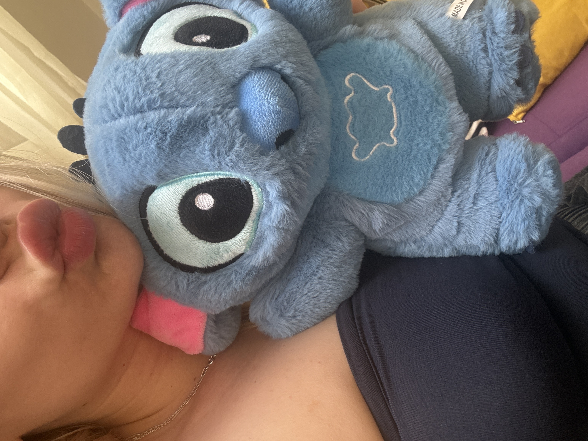 A woman is shown in a close-up, casual, personal photograph. She is fair-skinned with blonde hair visible in the lower left corner, with her face partially shown. The focus is on a plush toy of the Disney character Stitch, which is large and predominantly blue. The toy is positioned in the foreground, partially obscuring her face and resting on her chest. A pink tongue sticks out from the toy. The woman is wearing a dark blue top, and a thin silver necklace is visible on her neck. The background is out of focus, but shows a purple and yellow pillow. The photograph is taken indoors. There is no evidence to suggest the location is Sofia, Bulgaria.