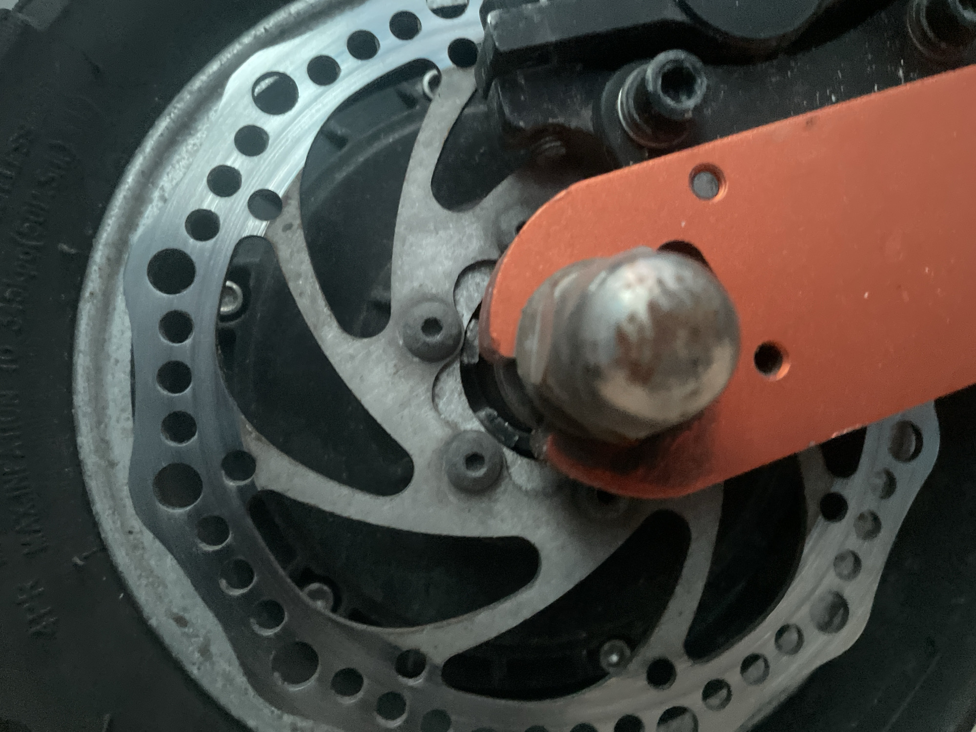 The media file shows a close-up, overhead view of a brake disc assembly on what appears to be a bicycle or scooter wheel. The subjects are the mechanical components of the braking system.
The scene is a detailed, possibly outdoor setting given the presence of a tire with visible text, though the immediate environment is not discernible. The activity is static, focusing on the stationary components of the brake.
Notable details include the perforated metal brake disc, a rusty silver-colored metal caliper component, and a bright orange metal arm extending from the assembly. There are several bolts and screws visible, securing the brake components. The overall impression is of a functional but somewhat worn piece of machinery, possibly from a vehicle.
Visible text on the tire, in the lower left quadrant, reads "MAX. INFLATION TO 345KPA(50PSI)".
The image does not contain any people, and there are no clear indicators of time of day or weather. The location context is provided as Kołobrzeg, Poland, but this cannot be visually confirmed from the image itself.
