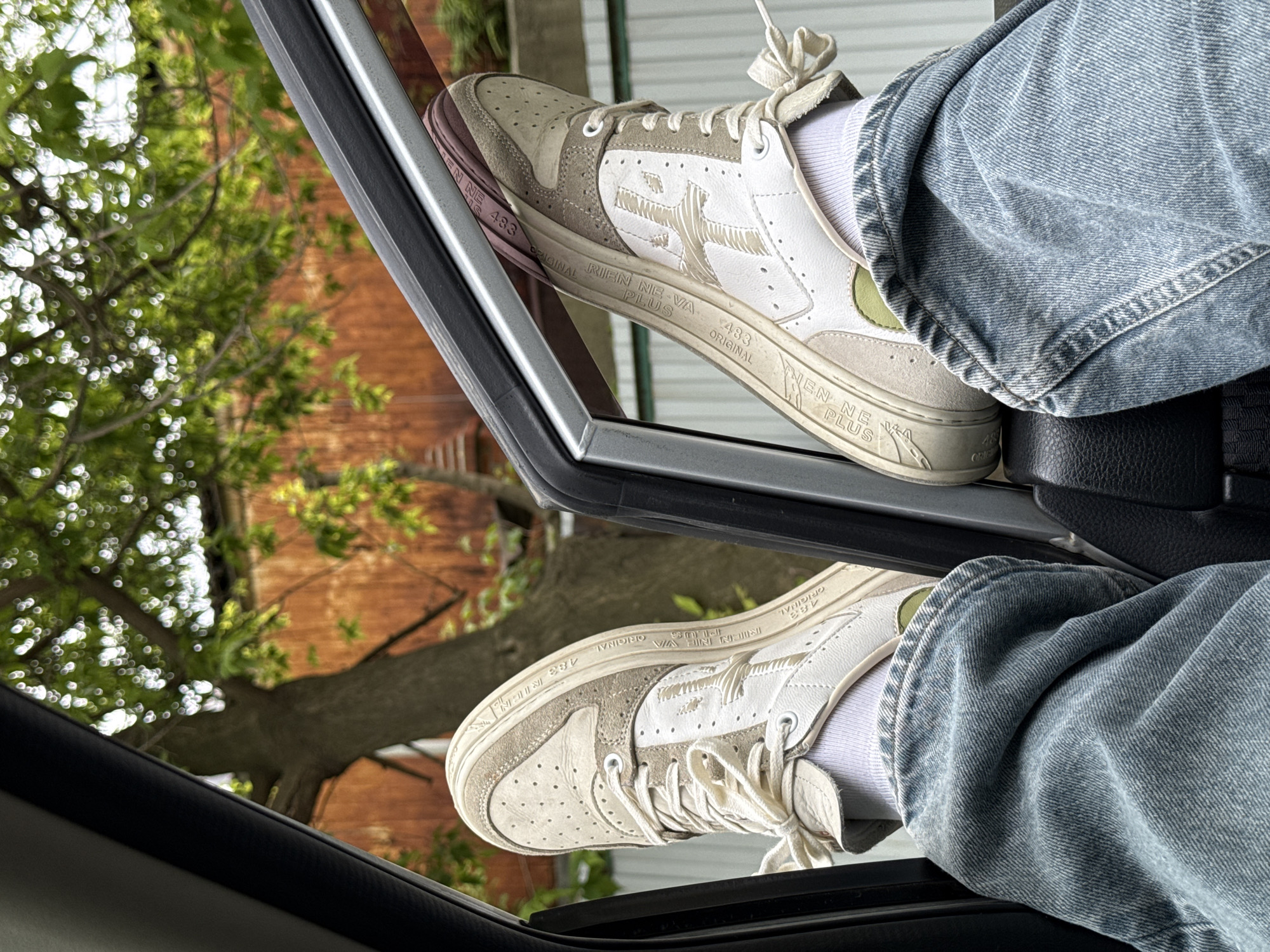 This vertical image captures a casual scene from inside a car, featuring a person's lower legs and feet. The individual is wearing light blue denim jeans and white socks, with their feet propped up on the frame of a partially open car window. They are wearing white and beige sneakers, which display the prominent text "RIEN NE VA PLUS" and "483 ORIGINAL" repeatedly along the sole and sides.
Through the window, a vibrant outdoor setting is visible. Lush green trees with abundant foliage fill the midground, with sunlight creating bright bokeh effects among the leaves. In the background, a building with reddish-brown brickwork can be seen. The natural lighting suggests it is daytime. The scene evokes a relaxed atmosphere, possibly during travel or a break, in an urban or suburban environment in Vladivostok, Russia. The car's black interior, including what appears to be a seat or armrest, is partially visible in the bottom right corner.