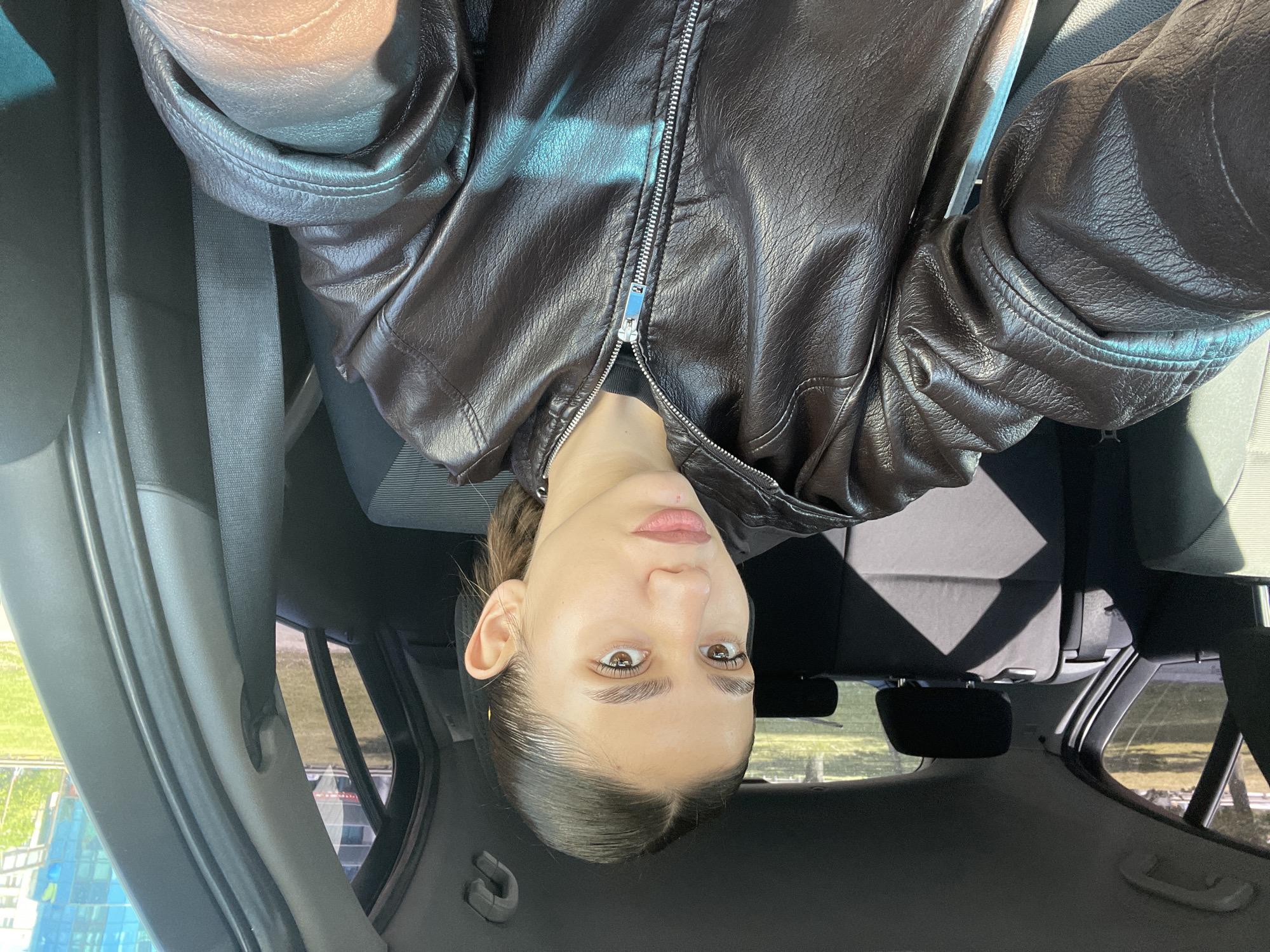 The image shows a person wearing a brown leather jacket with a silver zipper, seated in a car. The view is from an unusual angle, possibly taken from above and inverted, within the interior of the vehicle. The person is fair-skinned and has light brown eyes. The background visible through the car windows includes a city landscape. This photograph was taken in Sofia, Bulgaria. The photo shows parts of the car interior, including the headrest, seat and safety belt.