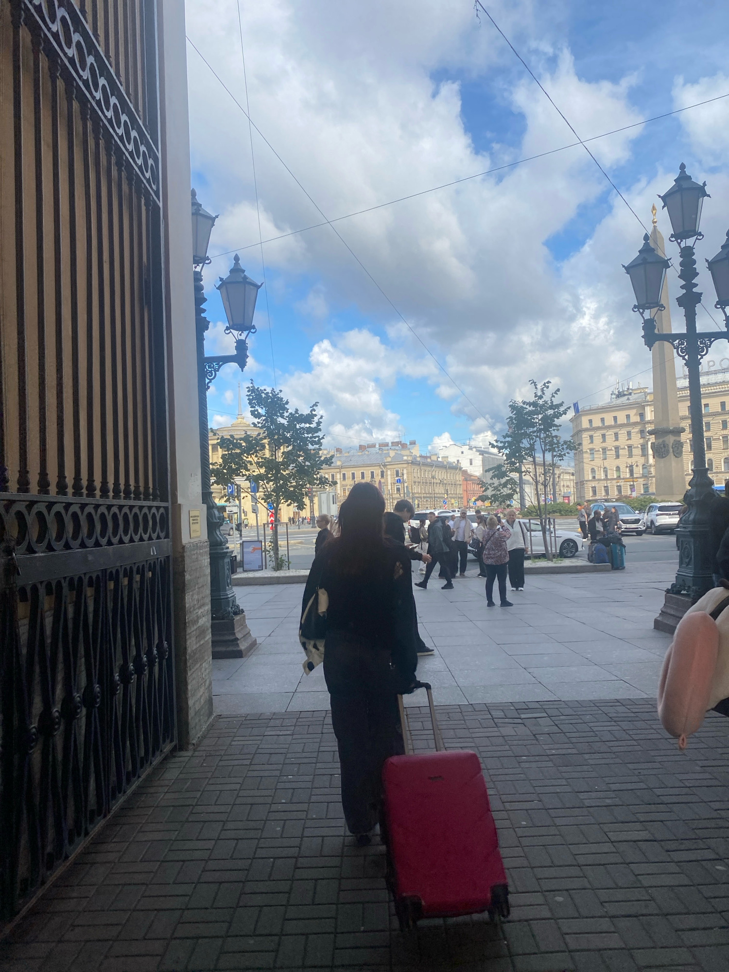 The image depicts a view from the entrance of a building in Saint Petersburg, Russia. The foreground shows a black wrought-iron gate and a brick pillar. A woman is seen from behind, walking away from the camera, pulling a red suitcase on wheels. She is dressed in dark clothing and carrying a bag over her shoulder. The background reveals a city street with pedestrians, buildings, trees, and street lamps. The sky is partly cloudy with blue patches. In the distance, an obelisk is visible. The street appears to be paved with rectangular stones.