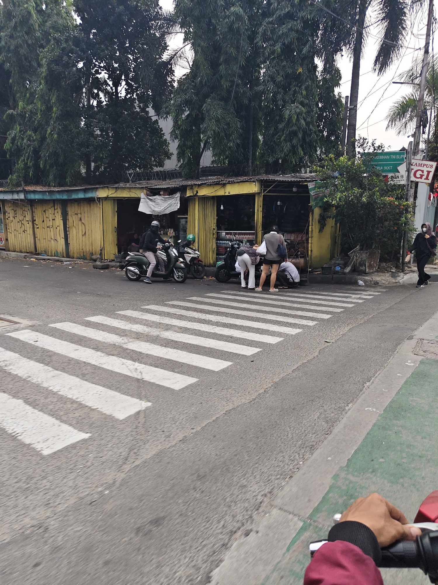 The image shows a street scene in West Jakarta, Indonesia, focusing on a pedestrian crossing and a small shop. In the foreground, a pedestrian crossing with white stripes is visible on the asphalt road. To the right of the crossing, a cyclist's hand wearing a burgundy jacket is holding a motorcycle handlebar. Beyond the crossing, a yellow building with an open storefront houses a shop, possibly a mechanic or a small convenience store. Several people are gathered near the shop. Two men and two women are interacting, with some of them bending down, suggesting they might be engaged in a transaction or repair. A green motorcycle is parked in front of the shop. Further down the street, another motorcycle and a woman in dark clothing are visible. The background is filled with lush green trees, and a street sign with Indonesian text is also present. The overall atmosphere suggests a typical day in an urban area of West Jakarta.