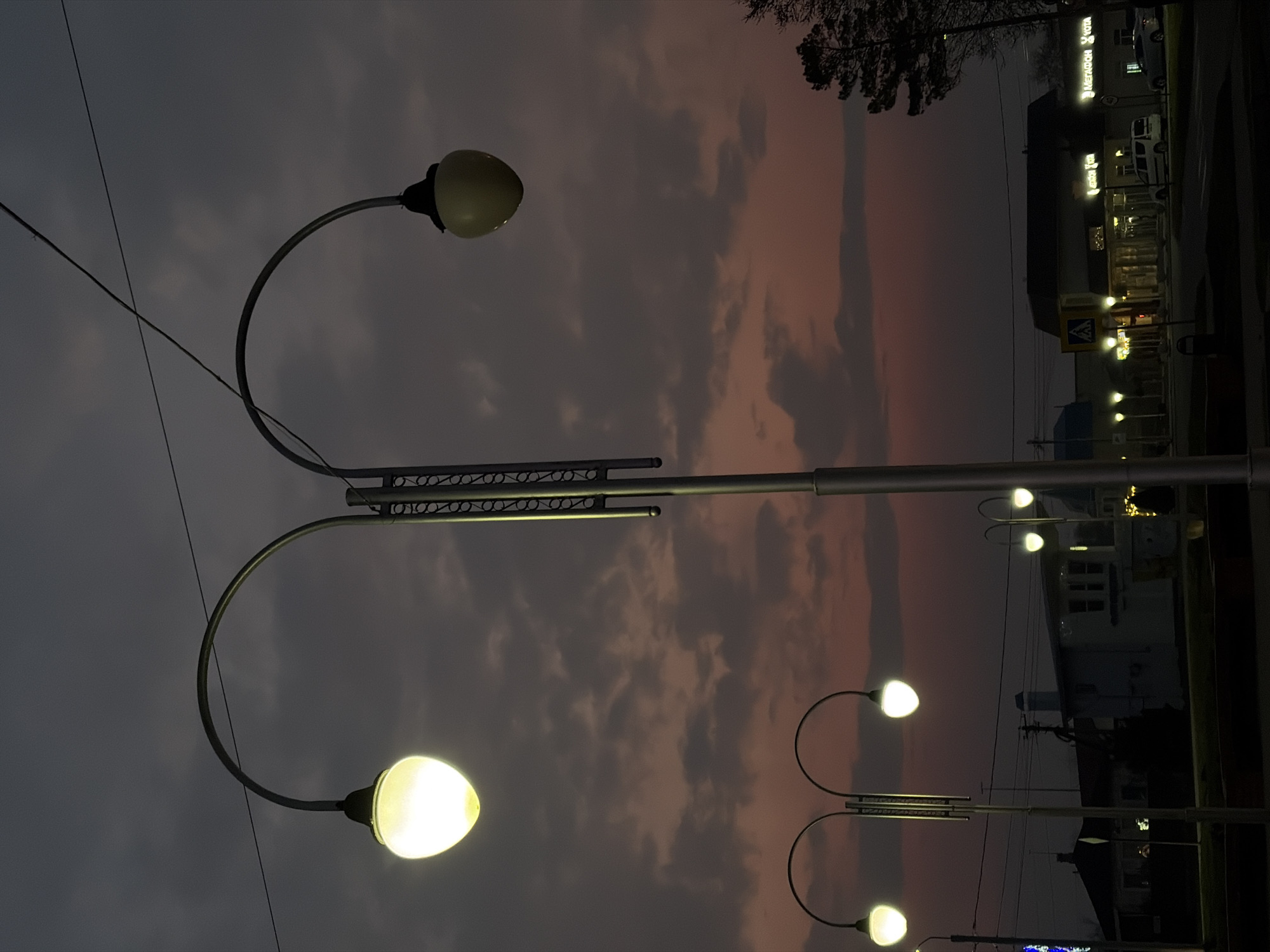 The image shows a street scene in Novopokrovskaya, Russia at dusk or night. There are streetlights in the foreground, with two prominent lamps illuminating the bottom of the frame. In the background, buildings with illuminated windows and signs are visible. The sky is a mix of dark gray and pink hues, suggesting a sunset or twilight. Power lines can be seen diagonally crossing the upper left corner of the frame.