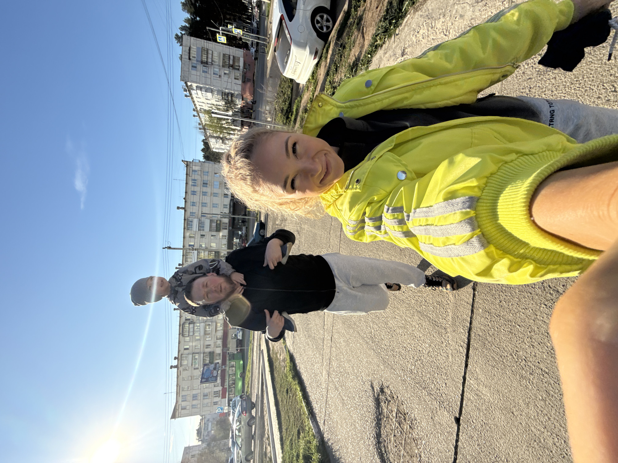 The image depicts a woman in a bright yellow jacket, holding a camera, presumably taking a selfie with two males and a child on a city street. One male is carrying the child on his shoulders. The group is standing on a paved road next to a grassy median. Buildings and parked cars are visible in the background.
Given the context of the city of Chaikovsky, Russia, and the architectural style of the buildings in the background, the location is likely a residential or commercial area within the city. Specific details that would pinpoint the exact location are not available from the image.