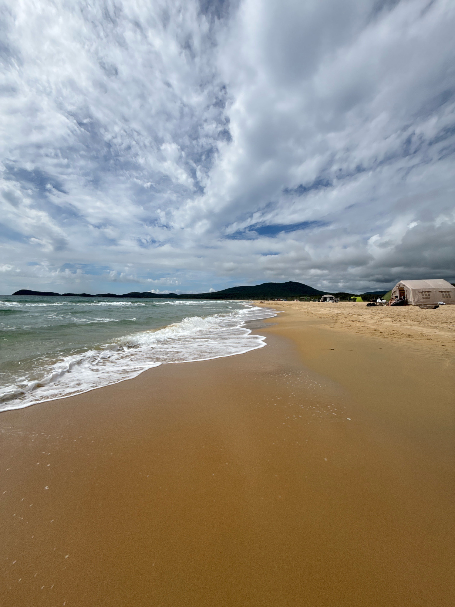 The image depicts a sandy beach with gentle waves lapping at the shore. The sky is filled with dramatic, textured clouds. In the background, a line of green hills or mountains borders the coastline. Several tents are set up on the beach, suggesting a camping or recreational activity. A few people are visible near the tents. The scene conveys a sense of natural beauty and outdoor leisure. This is not an image from Anna, Russia.