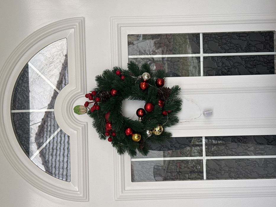 A white exterior door is depicted in Cibukovac, Serbia, adorned with a Christmas wreath. The door features a semicircular transom window above, containing multiple panes of textured glass, reflecting a portion of a tiled roof. Below the transom, two vertical rectangular panes of textured glass are present, separated by a white vertical mullion. A green artificial evergreen wreath, decorated with red berries, brown pinecones, red, gold, and silver spherical ornaments (including some bells), is centrally hung on the door. Small white LED fairy lights are integrated into the wreath, with a translucent white battery pack visible hanging by its wire below the wreath. The wreath is suspended from a small, light green, sprout-shaped hook positioned on the door directly below the arched transom moulding. No individuals are present, and no overt actions or interactions are occurring.