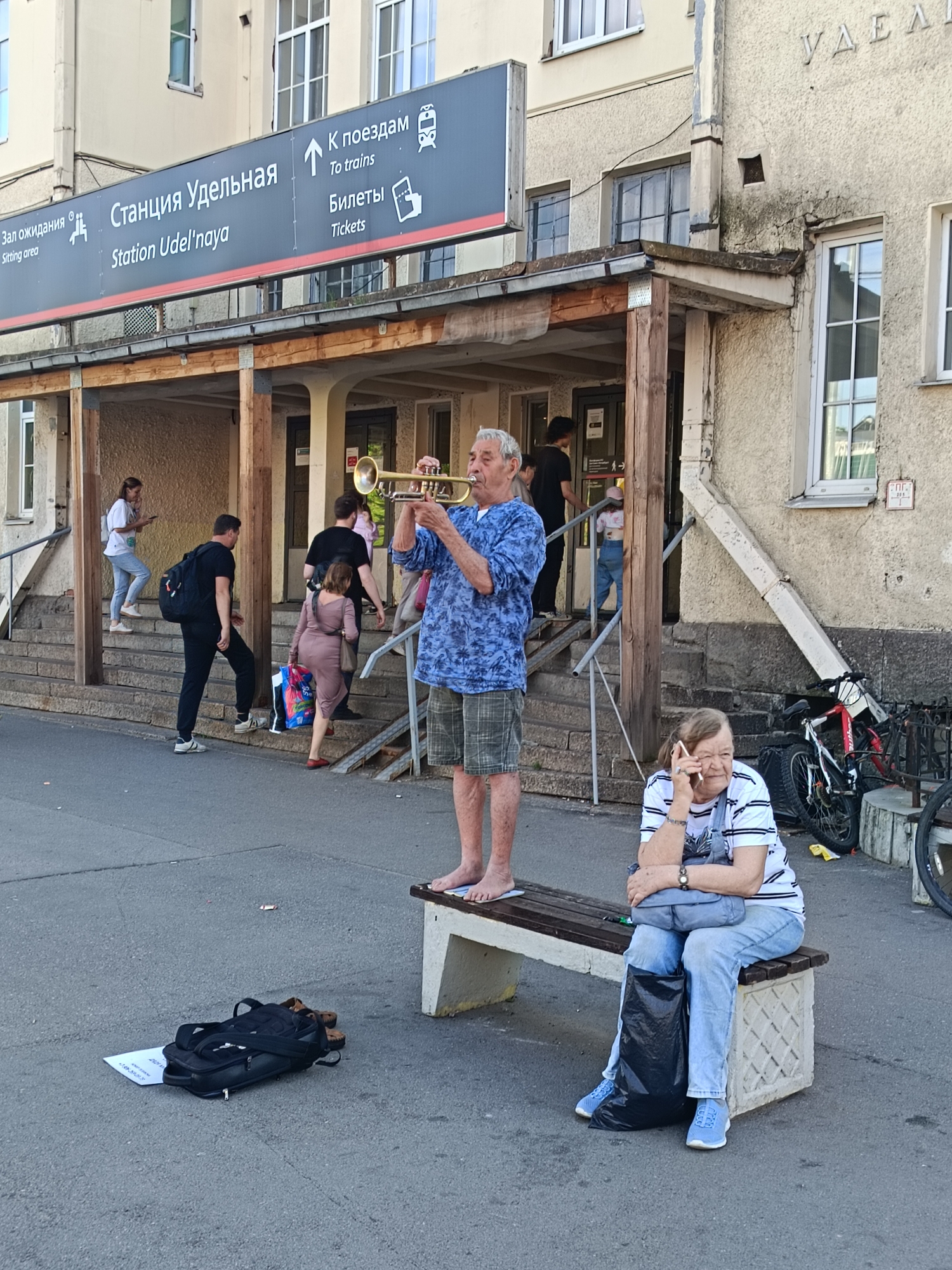 A man plays a trumpet in front of a train station building, likely located in Saint Petersburg, Russia, as indicated by the Russian signage that reads "Станция Удельная" and "К поездам," translating to "Udelnaya Station" and "To trains" respectively. The man, wearing a blue patterned shirt and plaid shorts, stands in the foreground, his bare feet visible. To his right, a woman sits on a bench, talking on her phone, dressed in a striped shirt and jeans, with a black trash bag beside her. Behind the trumpeter, several people ascend and descend stairs leading into the station. A black backpack and a piece of paper with writing are placed on the ground near the trumpeter's feet. Bicycles are parked in the background. The architecture of the station appears weathered, with a concrete facade and a covered entryway supported by wooden beams. The overall scene depicts a moment of public activity at a transit hub.