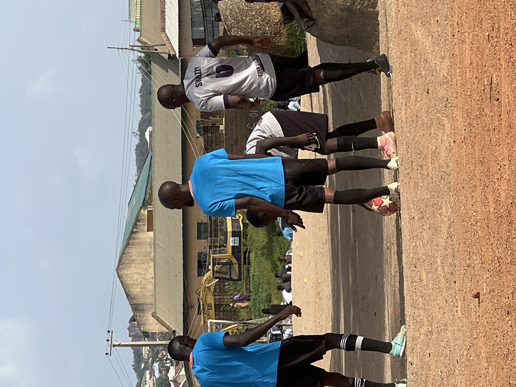 In the city of Jos, Nigeria, four Black males are engaged in a game of soccer on a dirt field. Three of the males are wearing blue t-shirts, while the fourth is wearing a gray t-shirt with the word "SWAZZY" printed on the back, along with the number "6". The field is surrounded by buildings and a yellow excavator. The sky is overcast.