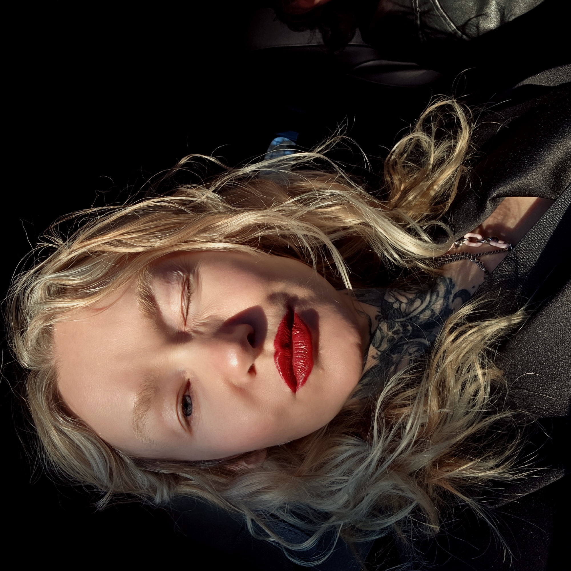 A fair-skinned person with blonde hair and prominent red lipstick is lying down on a black surface, potentially leather, with their head angled towards the top left of the frame. The person has tattoos on their neck and wrist, the wrist also adorned with a chain bracelet with pale pink links. The light is uneven, casting shadows across their face. There is no evidence to suggest the location is Saint Petersburg, Russia.