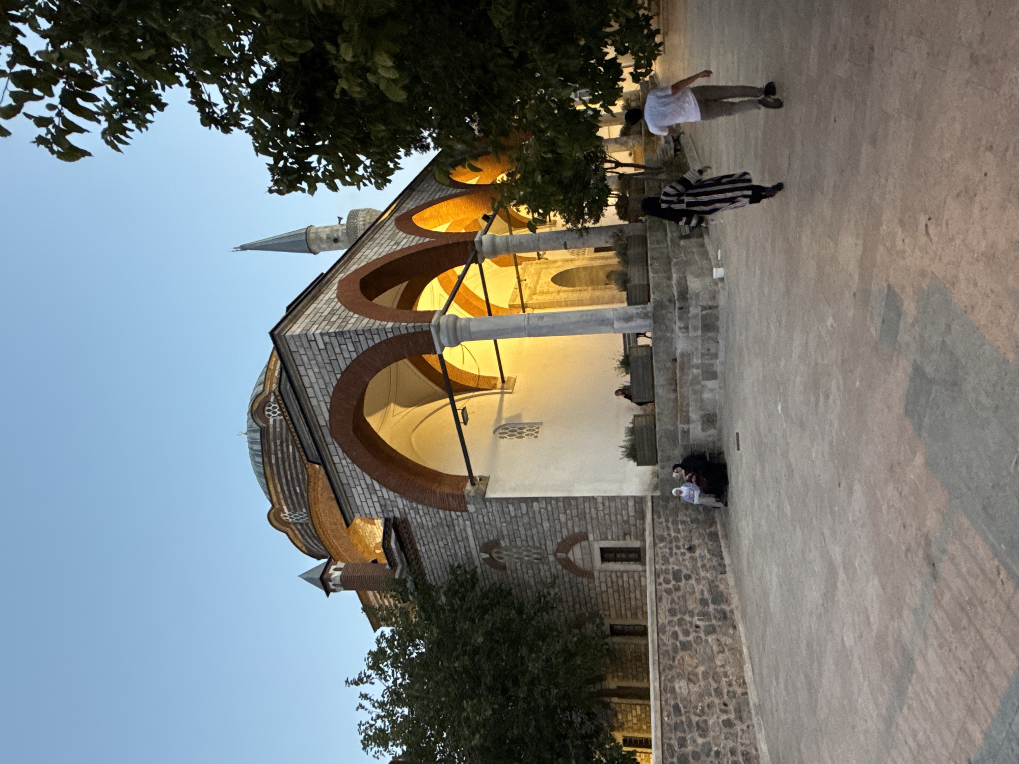 A mosque with a dome and a minaret is visible in the background of the image. The mosque is made of stone and brick, with arched doorways and windows. The exterior of the mosque is lit by warm artificial lights, casting a glow on the surrounding courtyard. A stone wall surrounds the courtyard, and several trees are planted within and around it, their leaves casting shadows on the ground.
In the foreground, a paved courtyard is visible. Two people, a woman in a striped dress and a man in a white t-shirt and grey pants, are walking away from the camera. A group of three women are sitting on a stone bench to the left of the image. They appear to be dressed in traditional Islamic clothing. The sky is a clear, pale blue, indicating that it is either early morning or late evening.
The location of this mosque is in the Üsküdar district of Istanbul, Turkey. It is likely the Mihrimah Sultan Mosque, built in the 16th century by the Ottoman architect Mimar Sinan. The mosque is one of the most famous landmarks in Üsküdar and is a popular tourist destination.