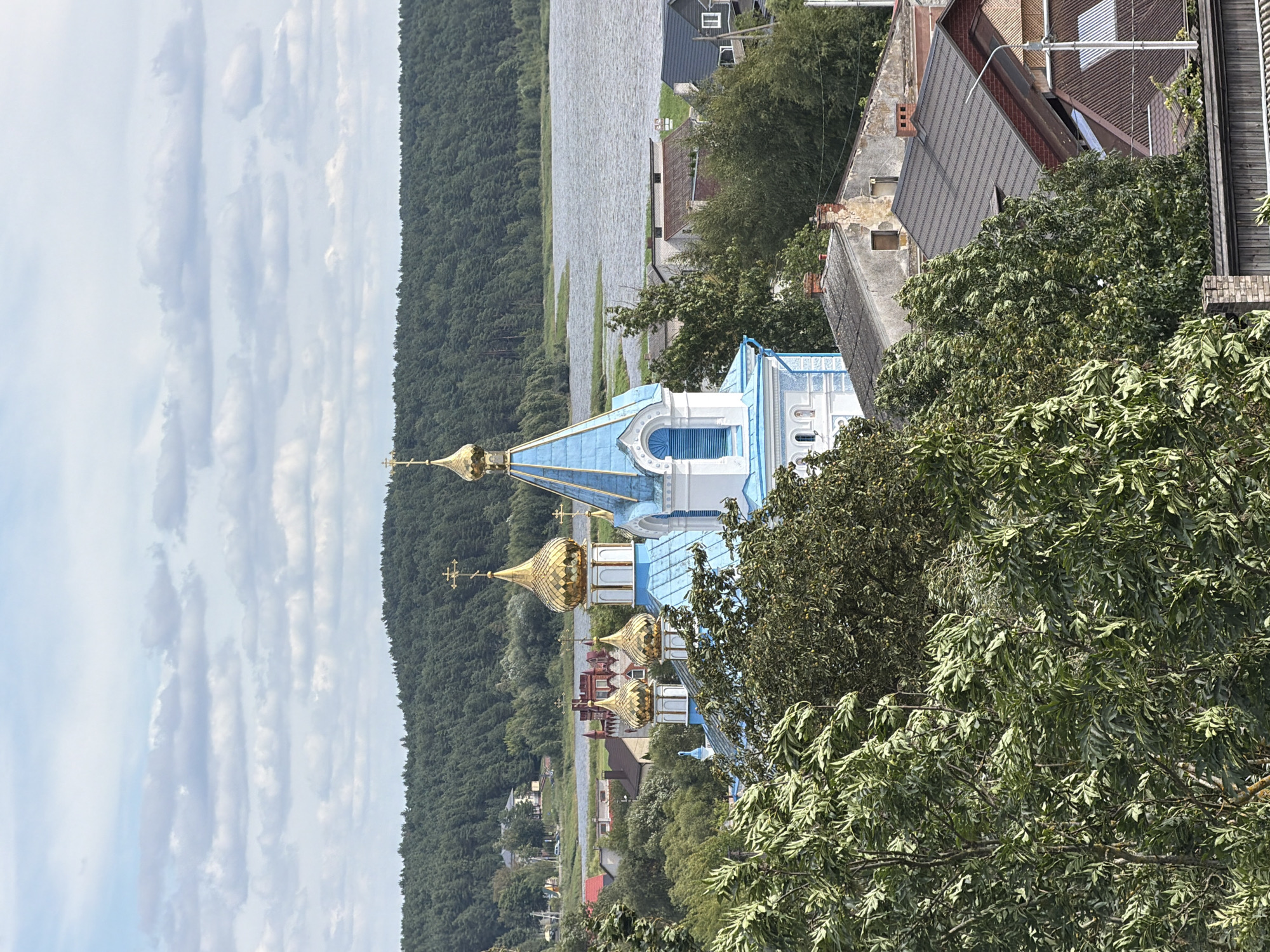 An outdoor, eye-level, long shot captures an Orthodox church with blue and white architecture and golden domes. The church is surrounded by trees, and in the background, a body of water is visible with a tree-covered hill beyond it. Several houses with dark roofs are scattered amongst the trees in the mid-ground. The sky is mostly cloudy with patches of blue.