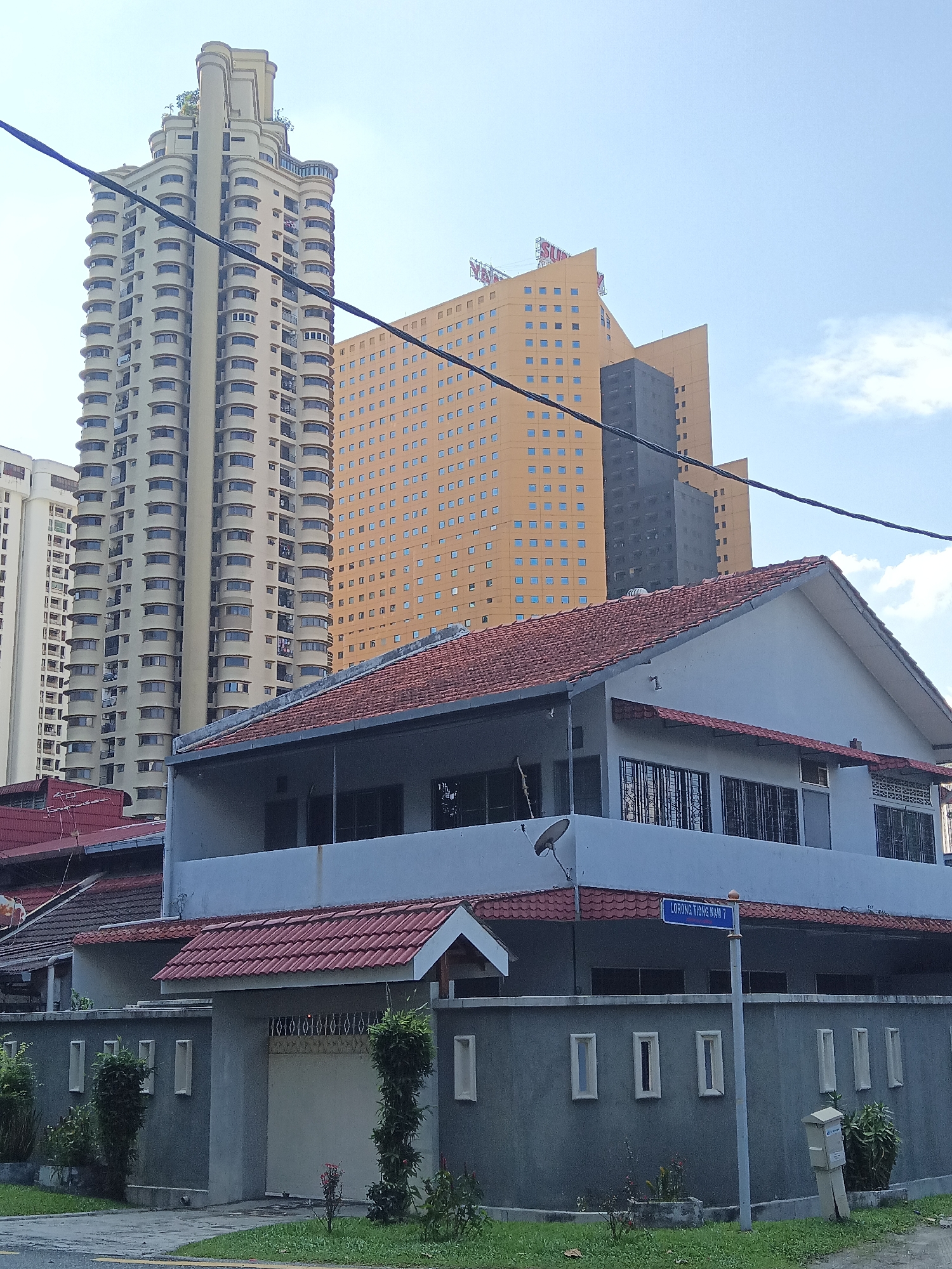 The image captures a street view in the Tiong Nam area of Kuala Lumpur, Malaysia, on a clear day with scattered clouds. In the foreground, a single-story house with a red tiled roof is prominent, featuring a grey concrete fence with rectangular openings and decorative elements. A blue street sign indicates "LORONG TIONG NAM 7." To the left of the house, the red tiled roofs of adjacent houses are visible. Behind the foreground house, a taller, modern residential building with a beige facade and numerous balconies stands out. Further in the background, a large, orange-colored hotel or office building with a grid of square windows dominates the skyline. To the right of the orange building, a darker, narrower tower is visible. Power lines are strung across the upper portion of the image. The street in front of the house is paved, with a strip of green grass and some landscaping. No people are visible in the scene.