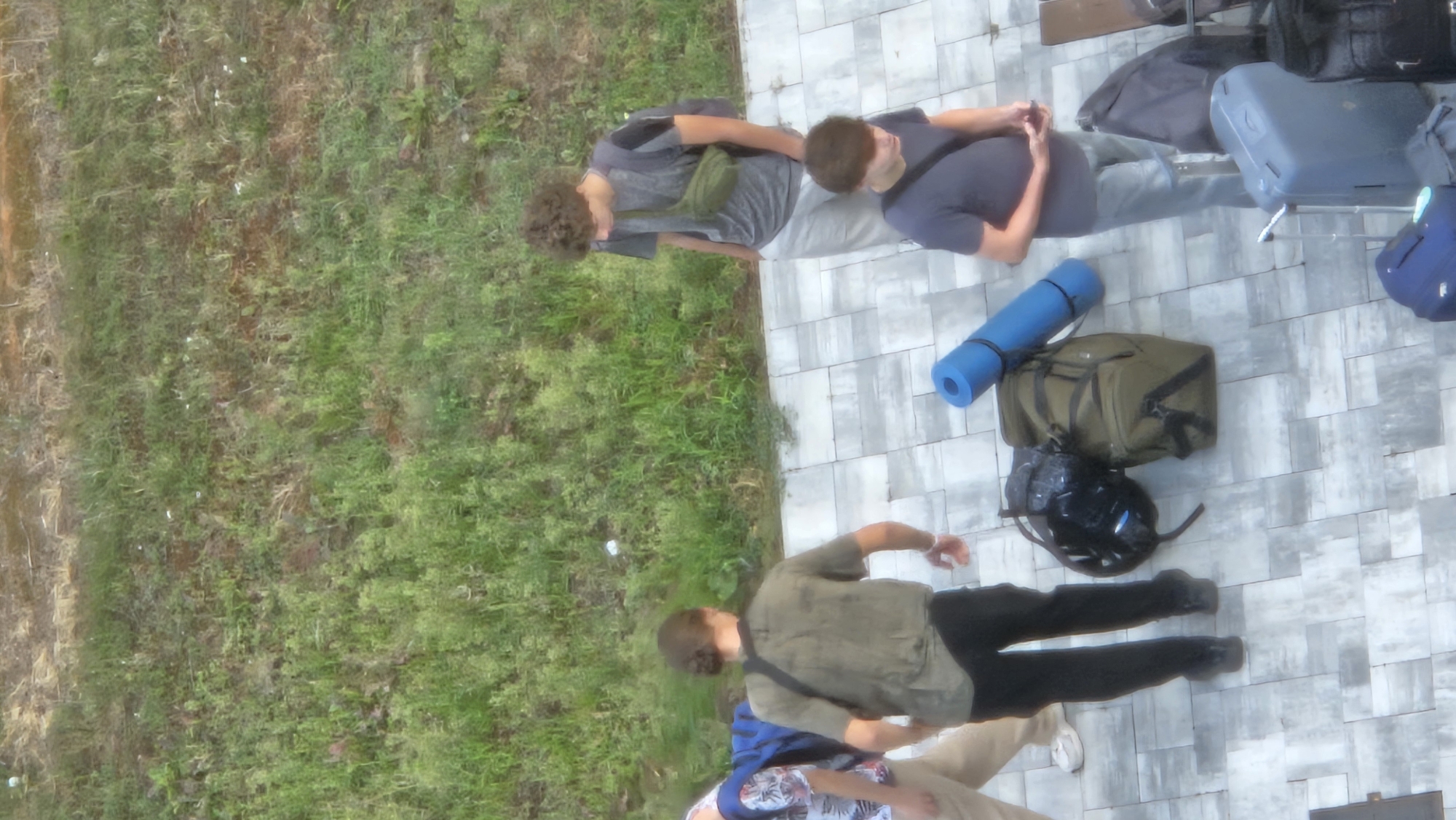This is a high-angle, outdoor, candid photograph of a group of young people and their luggage on a paved area next to a grassy slope. The scene appears to be in the city of Prešov, Slovakia, possibly near a transportation hub or a park.
In the foreground and center, there are several pieces of luggage. A large olive green duffel bag is placed on the ground, with a bright blue rolled-up sleeping mat strapped across it. To the left of the duffel bag, a black helmet or round bag is visible. Further to the right, a grey rolling suitcase with its handle extended stands upright, and next to it, a dark blue rolling suitcase. Another dark bag is partially visible to the right of the grey suitcase.
There are at least four young men in the image.
The leftmost person, partially visible from the waist down, wears a floral print shirt and light beige pants with white sneakers. They are walking away from the camera.
To their right, a young man in a dark olive green short-sleeved shirt and black pants is standing and looking downwards, his right arm bent at the elbow. He has a dark strap across his torso.
A young man with curly brown hair, wearing a grey t-shirt and light grey pants, stands facing slightly away from the camera, looking downwards. He has a green fanny pack worn across his chest and a backpack on his back.
In the bottom right of the frame, another young man in a dark blue t-shirt and light grey pants is visible from the chest down, holding a mobile phone in his right hand. He also has a dark strap across his torso.
The environment consists of a paved area made of large rectangular tiles with a light grey and white pattern, adjacent to a lush green grassy slope covered with various types of vegetation. The lighting suggests it is daytime. The overall atmosphere appears casual, possibly indicating travel or outdoor activities.