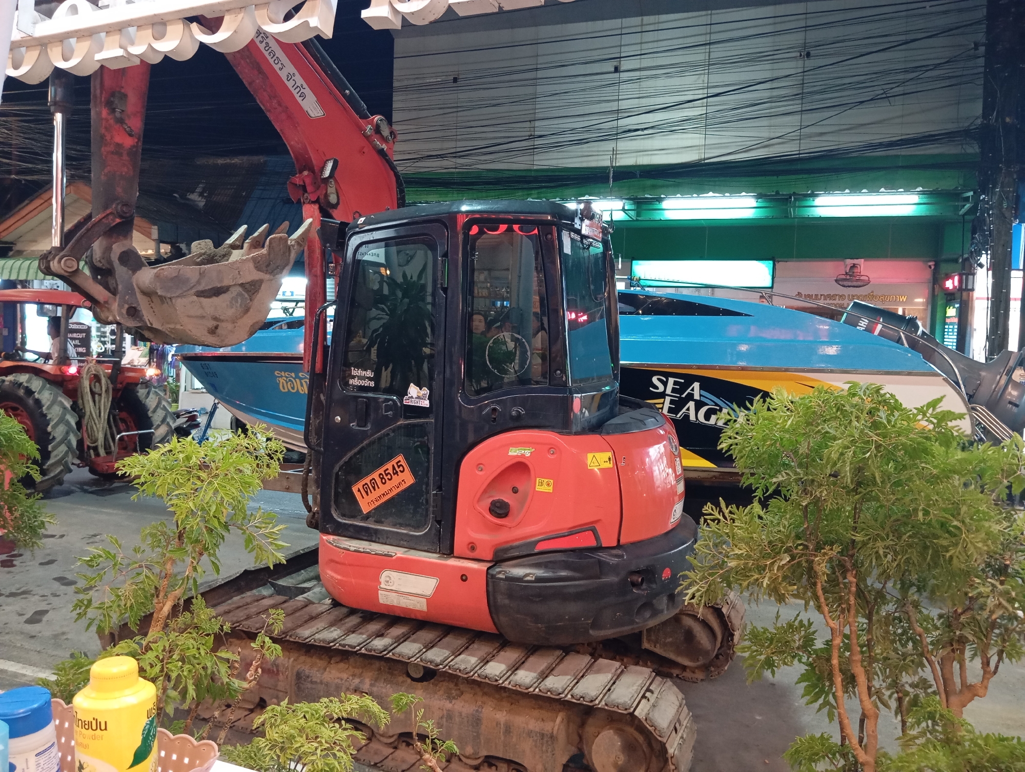 In Karon, Thailand, an orange and black excavator is parked on a grey paved surface. The excavator's cab is visible, with windows reflecting the surroundings. A red boom extends upwards from the excavator, holding a metal bucket filled with dirt. Behind the excavator, a blue and yellow boat with "SEA EAGLE" printed on its side is parked. The boat has an outboard motor. To the left of the excavator, the rear tire of a red tractor is visible. Green leafy plants are placed in front of the excavator and to its right, obscuring parts of the tracks. Overhead, a network of wires is visible against a dark background. A white awning with decorative white shapes is present at the top of the image. A yellow powder container with Thai writing is on a small table in the foreground. Several stickers are visible on the excavator, including one with Thai writing and a license plate number.