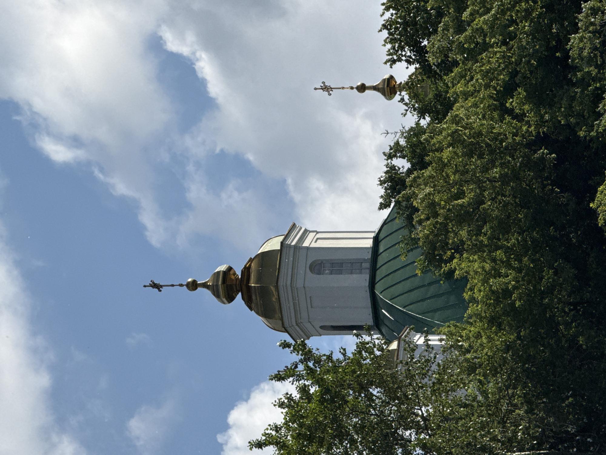 The image captures the upper portion of a church, likely located in the city of Perm, Russia. The prominent feature is a white octagonal bell tower topped with a golden dome and a cross. Adjacent to it, a large green dome of the church is visible, also crowned with a golden cross. The structures are partially obscured by lush green trees in the foreground. The sky above is a mix of blue and white clouds, suggesting daylight. The scene is devoid of any people or ongoing activities.