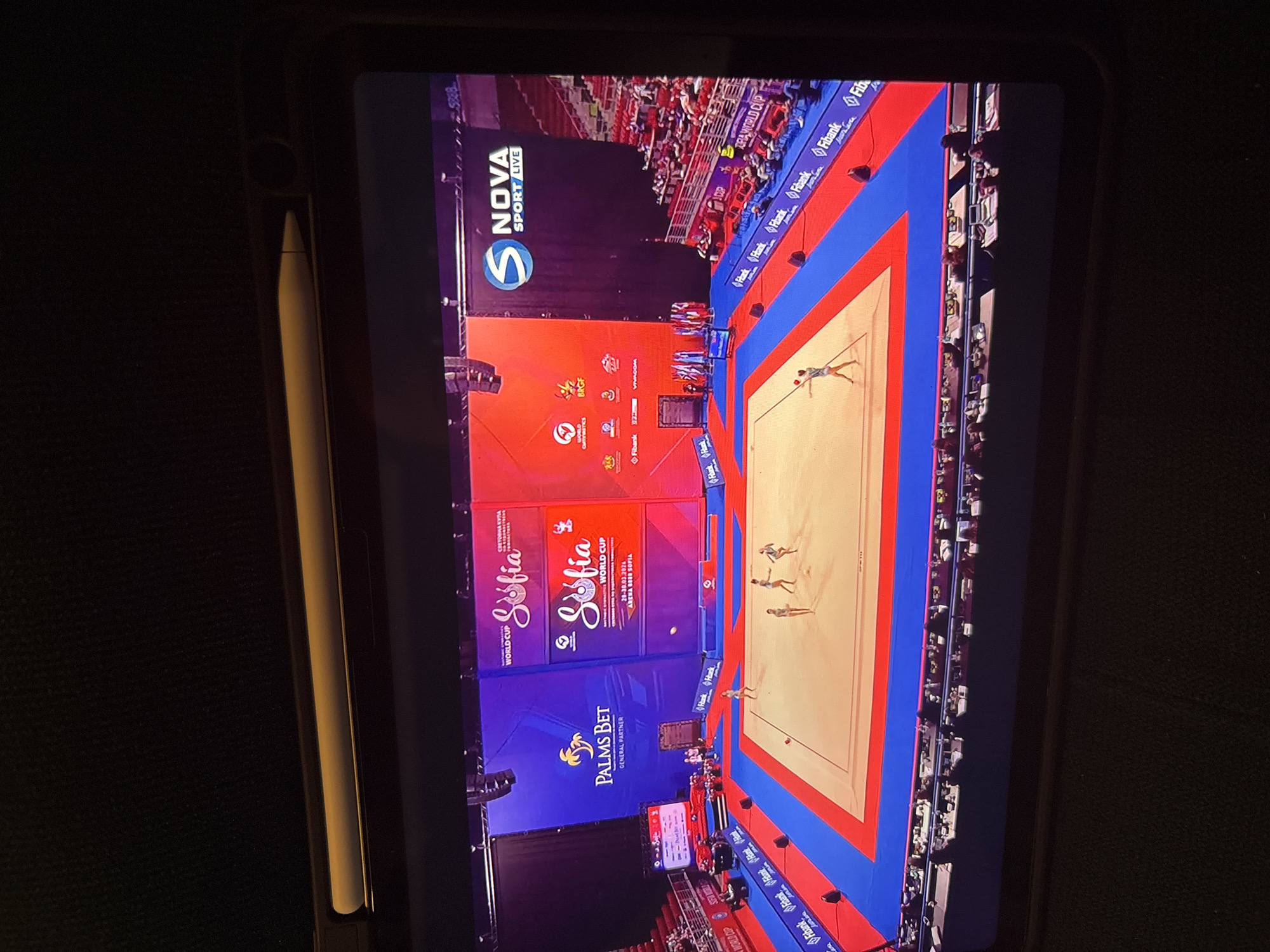 The image shows a tablet displaying a broadcast of rhythmic gymnastics competition in Sofia, Bulgaria. A gymnastics floor exercise performance is in progress on the screen. Banners behind the competition area show "Sofia World Cup," the dates 28-30.03.2024, "Arena Sofia," and sponsor logos including "Fibank" and "Palms Bet." There are spectators in the stands. An Apple Pencil is attached to the side of the tablet. The broadcast is a NOVA Sport live event.