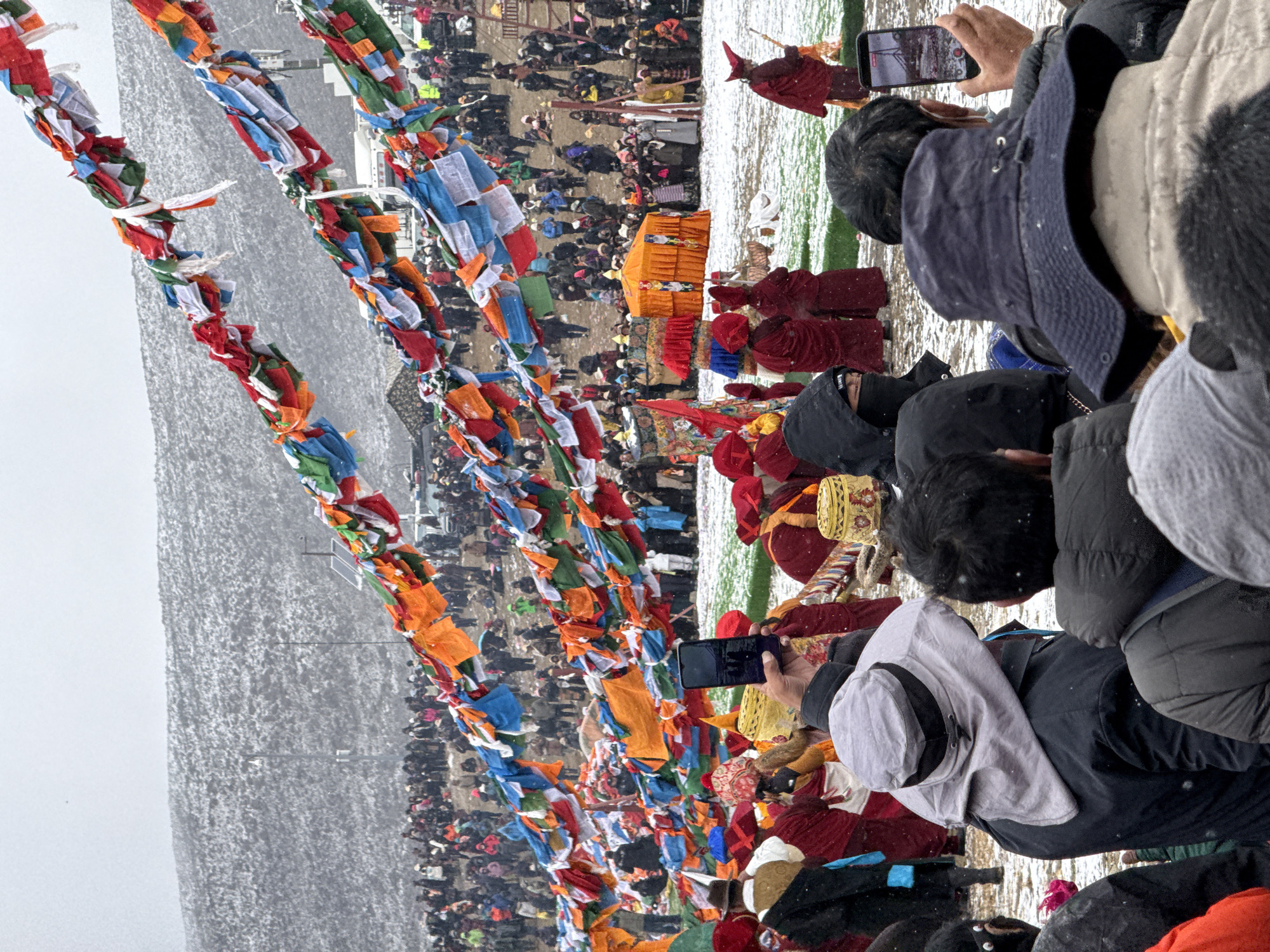 The image captures a crowded scene in what appears to be Parga Town, China. Numerous people are gathered on a snow-covered ground. Many individuals are dressed in what looks like monastic robes. Prayer flags with a multitude of colors are strung across the scene. Some attendees are using their phones to document the event. A raised platform decorated in orange and red fabric is visible among the crowd, seemingly indicating a focal point for the assembly. The backdrop consists of a snow-covered landscape under a cloudy sky.