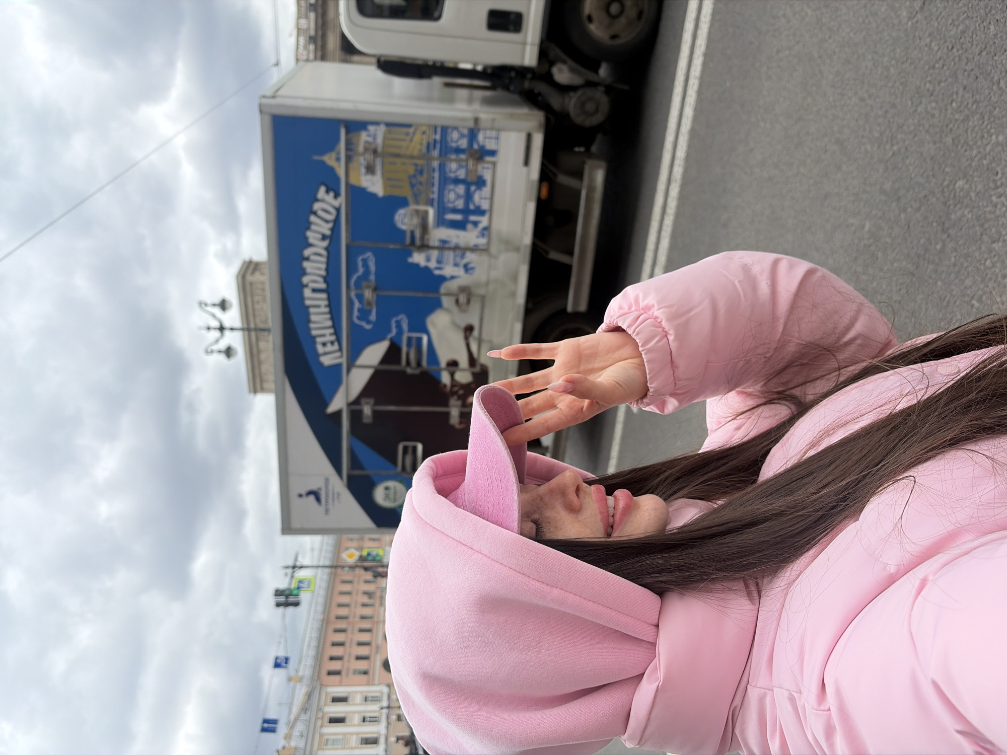 This image captures a vibrant street scene in Saint Petersburg, Russia, centered around a person posing playfully. The main subject is a young person, likely female, with long, dark hair, seen in profile. She is wearing a bright pink hooded sweatshirt, with the hood up, and a matching pink puffer jacket. Her head is tilted upwards, revealing a broad smile on her face, and her left hand is raised near her temple, forming a "peace" or "V" sign.
In the background, a white delivery truck is prominently featured on an asphalt street with white lane markings. The truck's side is adorned with a large blue graphic that includes the Cyrillic text "ЛЕНИНГРАДСКОЕ" (Leningradskoye), which directly references Leningrad, the former name of Saint Petersburg. Below the text, an illustration resembling a grand historical building or cathedral, characteristic of Saint Petersburg's architecture, is visible.
Beyond the truck, other elements of the urban environment are discernible, including classic-style buildings with numerous windows, traditional street lamps, and a traffic light. The sky above is completely overcast with thick grey clouds, indicating a cloudy daytime setting. The person's joyful expression and casual pose create a lively contrast against the historical and industrial backdrop of the city street.