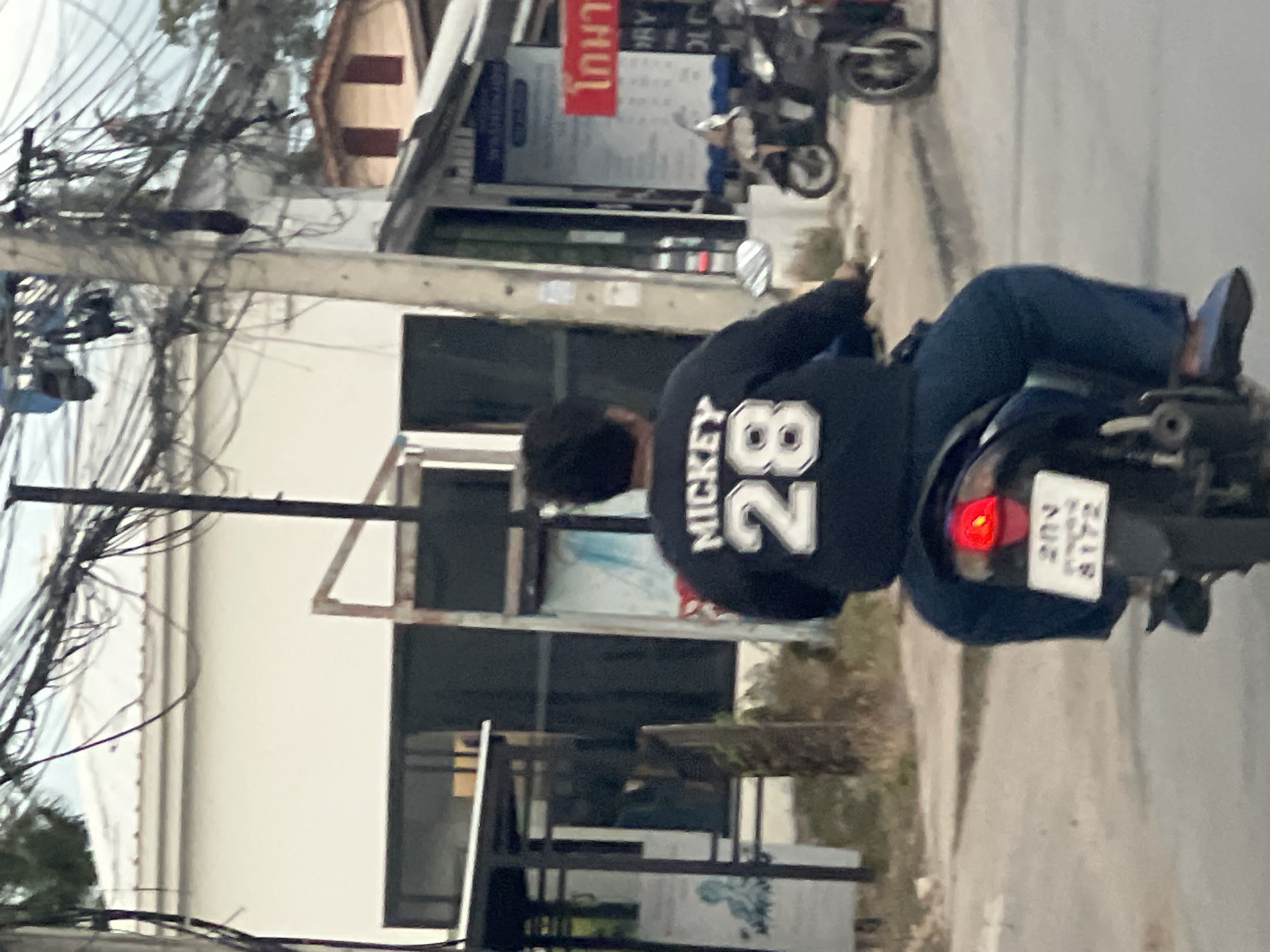 This image shows a person riding a motorcycle in an outdoor, urban setting. The rider is wearing a dark blue long-sleeved shirt with "MICKEY 28" printed in white on the back. They are also wearing blue pants. The motorcycle has a license plate with Thai characters and numbers that read "20V 8172". The background shows buildings, power lines, and a street. The sky is overcast, suggesting it might be cloudy or nearing dusk. The scene appears to be a street in a town or city in Ko Samui District, Thailand, given the presence of Thai text on the license plate and the architectural style of the buildings. The overall mood of the image is neutral, focusing on the mundane activity of riding a motorcycle.
