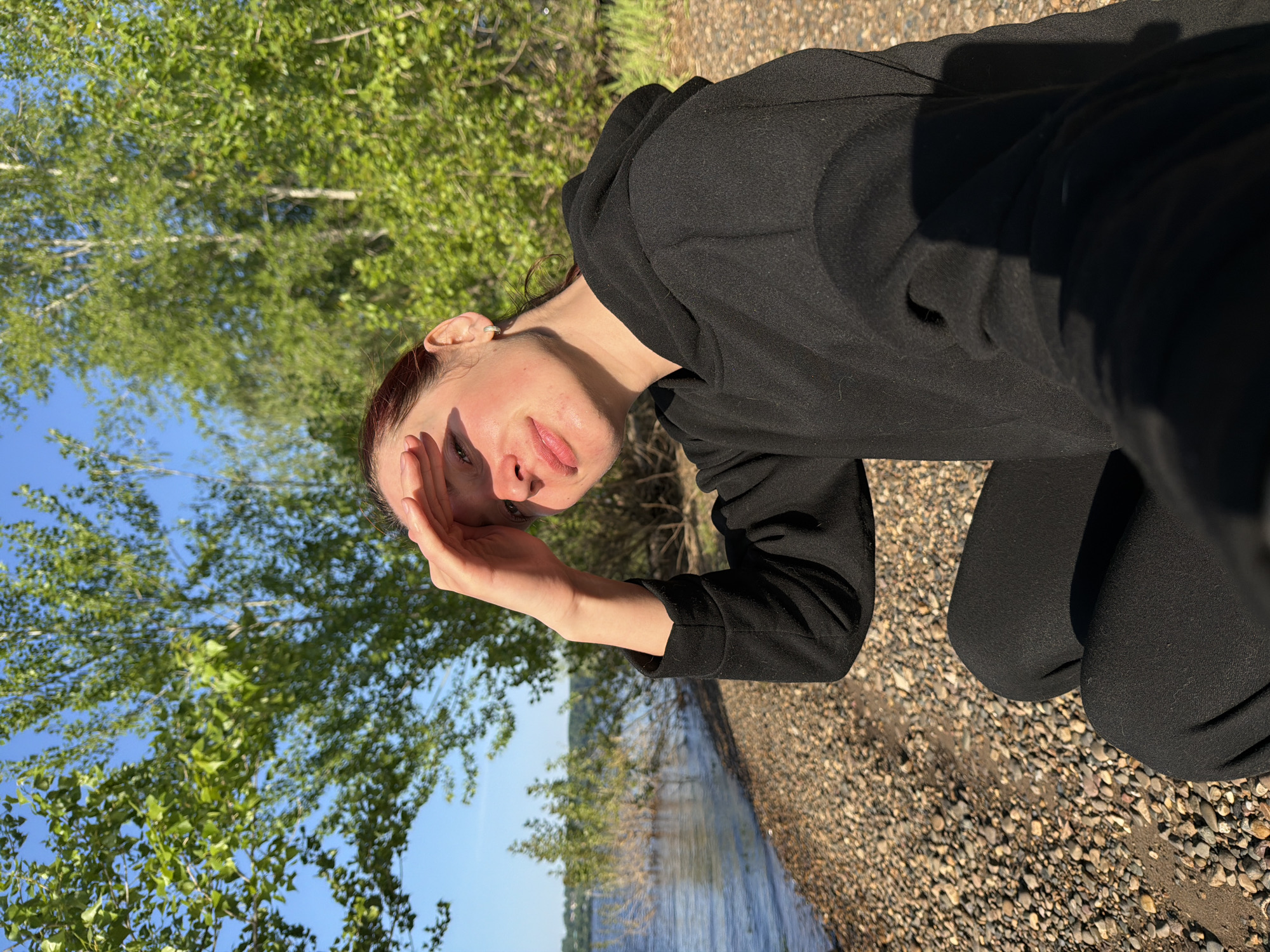 A person wearing a black hoodie and black pants kneels on a gravel beach, shielding their eyes with their left hand. Behind the person is a body of water, possibly a river or lake, with trees lining the opposite shore. A lush green tree is on the left side of the frame, and the sky is partially visible as blue. The location is Noviy, Russia.