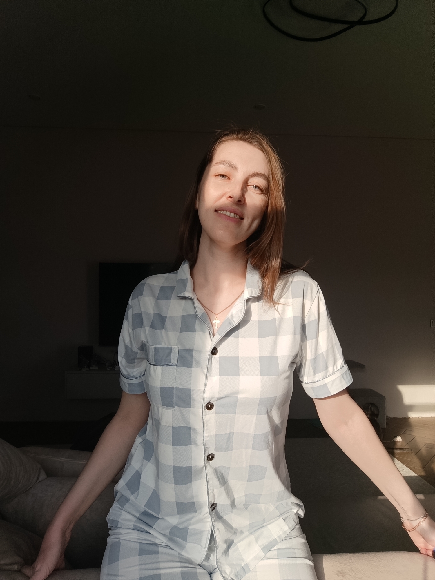 A woman with light skin and brown hair is sitting indoors, wearing a light blue and white checkered pajama set and a gold cross necklace. Her arms are outstretched. Behind her is a darkened room with a television and furniture visible in the background. Sunlight streams into the room from the right, casting shadows on the floor. The location is Chaikovsky, Russia.