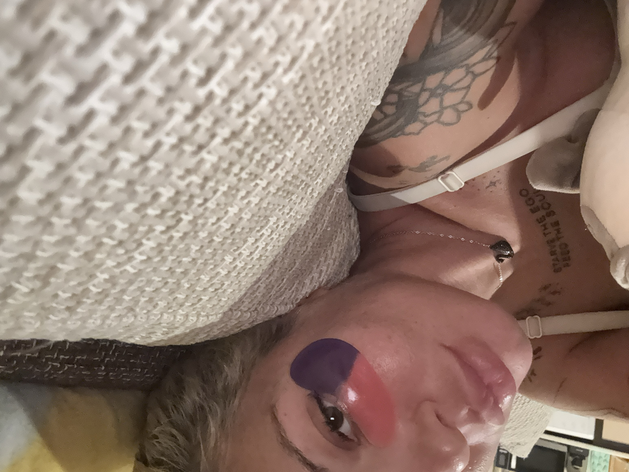 The image shows a close-up of a person's face, focusing on their eye and lips. The person has fair skin and is wearing eye patches under their eyes, one red and one purple. Their lips appear glossy and slightly parted. There are tattoos visible on the person's shoulder and neck. The person is lying down, with part of their face resting on a textured, light-colored surface, possibly a couch or pillow. The background is out of focus, showing glimpses of what appears to be a room with some furniture. The image does not provide any information about the location within the city of Plovdiv, Bulgaria.