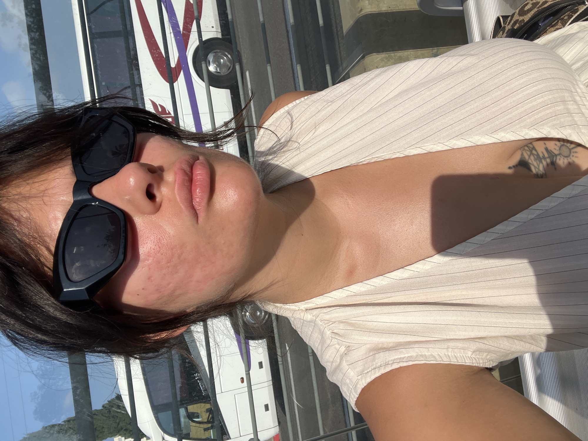 A woman wearing sunglasses and a sleeveless white top with pinstripes is seen from the chest up, in a selfie. She has dark hair pulled back and is looking towards the camera with her lips slightly parted. Redness is visible on her cheeks. In the background, through what appears to be a window, a white bus with purple and red stripes is seen driving on a road. The scene is outdoors, possibly on a sunny day, with a blue sky visible. The location is identified as Kiryat Motzkin, Israel.