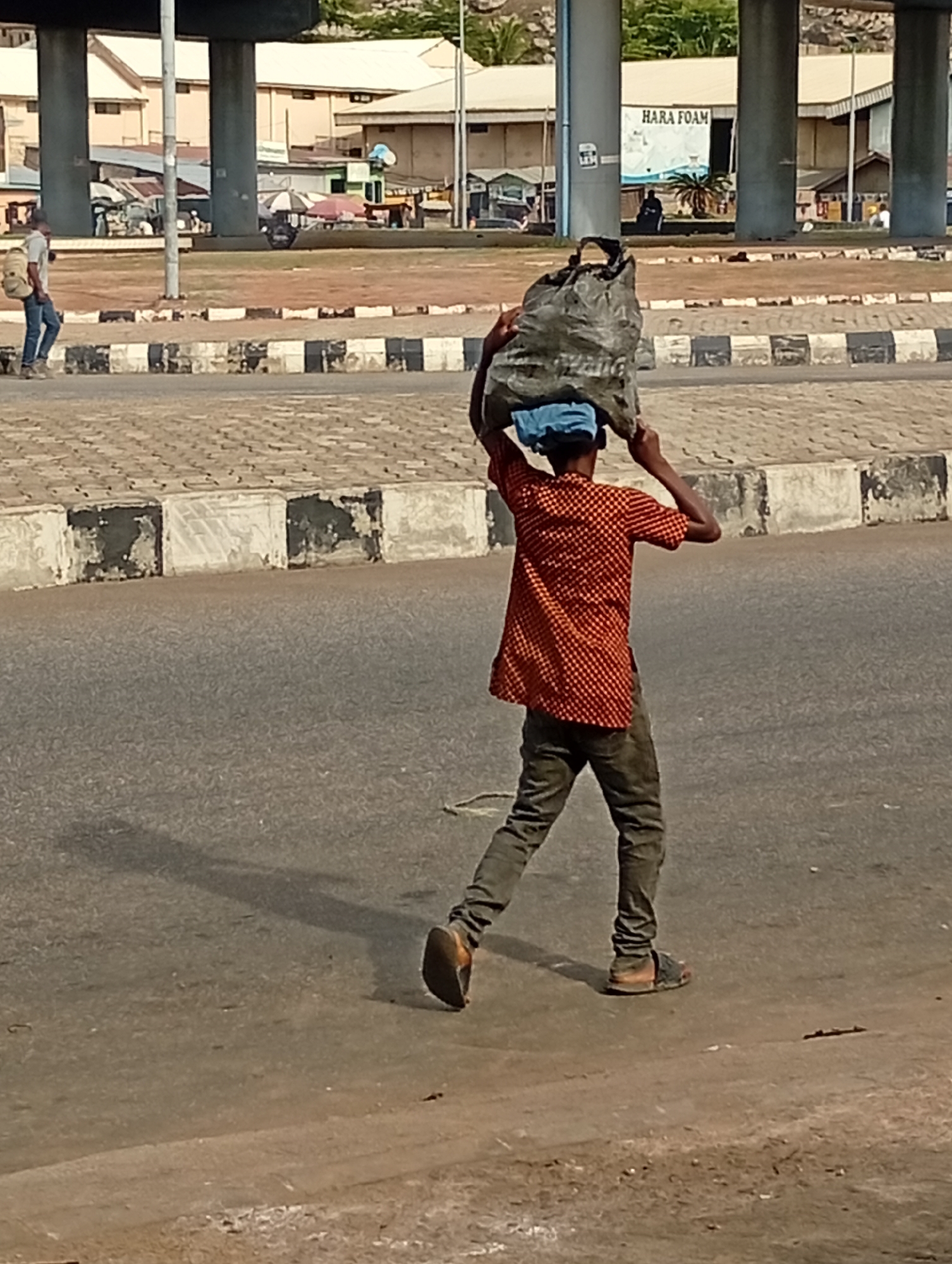 A person wearing a red checkered shirt, blue head covering, gray pants, and sandals is carrying a large gray bag on their head while walking on a paved road in Lokoja, Nigeria. The road is bordered by a black and white checkered barrier. In the background, under a concrete overpass, are buildings, including one with a sign reading "HARA FOAM." A man in a gray shirt and jeans walks in the background on the far side of the barrier. The scene is captured in daylight, and the person carrying the bag casts a long shadow on the road.