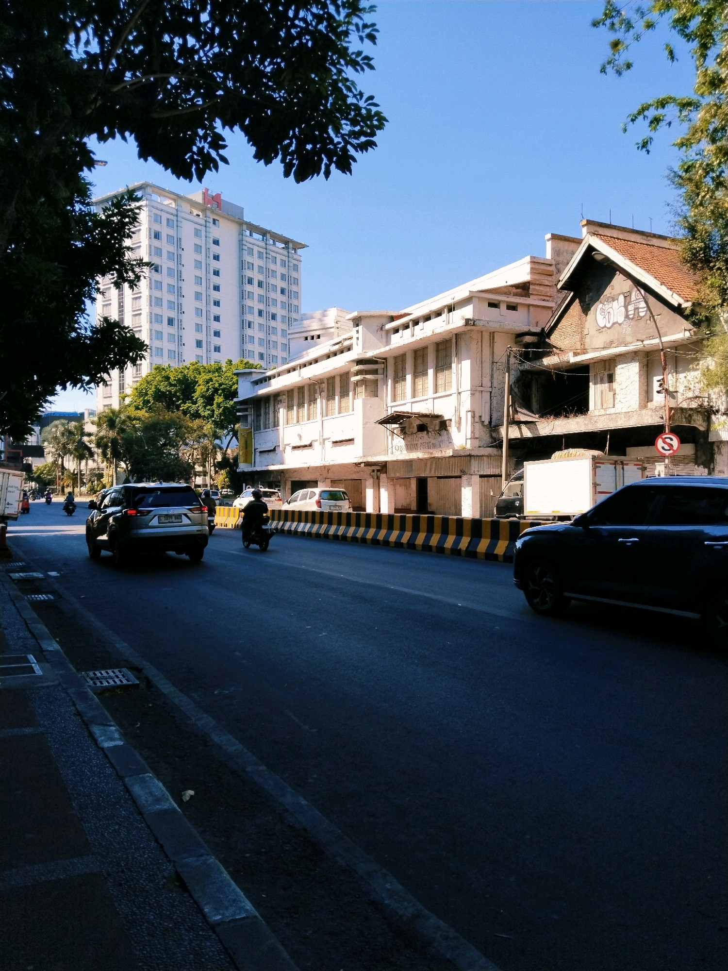 This image depicts a street scene in Surabaya, Indonesia, likely during daylight hours with clear skies. The foreground shows a paved road with a curb and sidewalk. A dark-colored SUV is driving away from the camera on the right side of the road, and a motorcycle rider is also on the road.
In the mid-ground, there is a white truck with a blank advertisement panel and a yellow and black striped barrier separating the road from the sidewalk. Several buildings line the street. On the left, a tall, white high-rise building with many windows dominates the background. In the center and right of the mid-ground, older, multi-story buildings with white facades and visible windows are present. Some of these buildings show signs of age and wear, with graffiti visible on one structure. Trees are scattered throughout the scene, with a large tree with dark leaves in the upper left corner casting a shadow. The overall impression is of an urban environment with a mix of modern and older architecture.