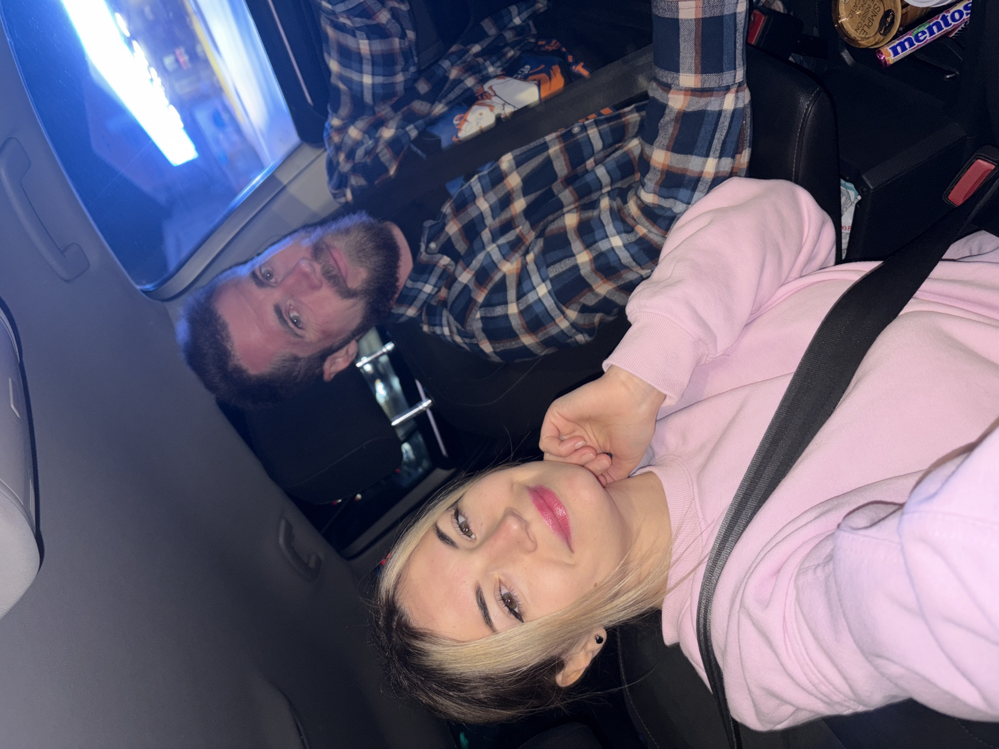 Inside a car, a woman and a man are seated with seatbelts fastened. The woman, positioned in the foreground, has fair skin, blonde streaks in her dark hair, and is wearing a pink sweater. She has pink lipstick on and is holding her hand near her face. Behind her, a man with a beard is wearing a blue and white plaid shirt and also has his seatbelt on. A window is visible in the upper left, reflecting city lights. In the upper right corner, items like a "mentos" package and a can are visible. The environment suggests they are in a vehicle, possibly driving through a city at night. The precise location in Budva, Montenegro is not discernible from the image.