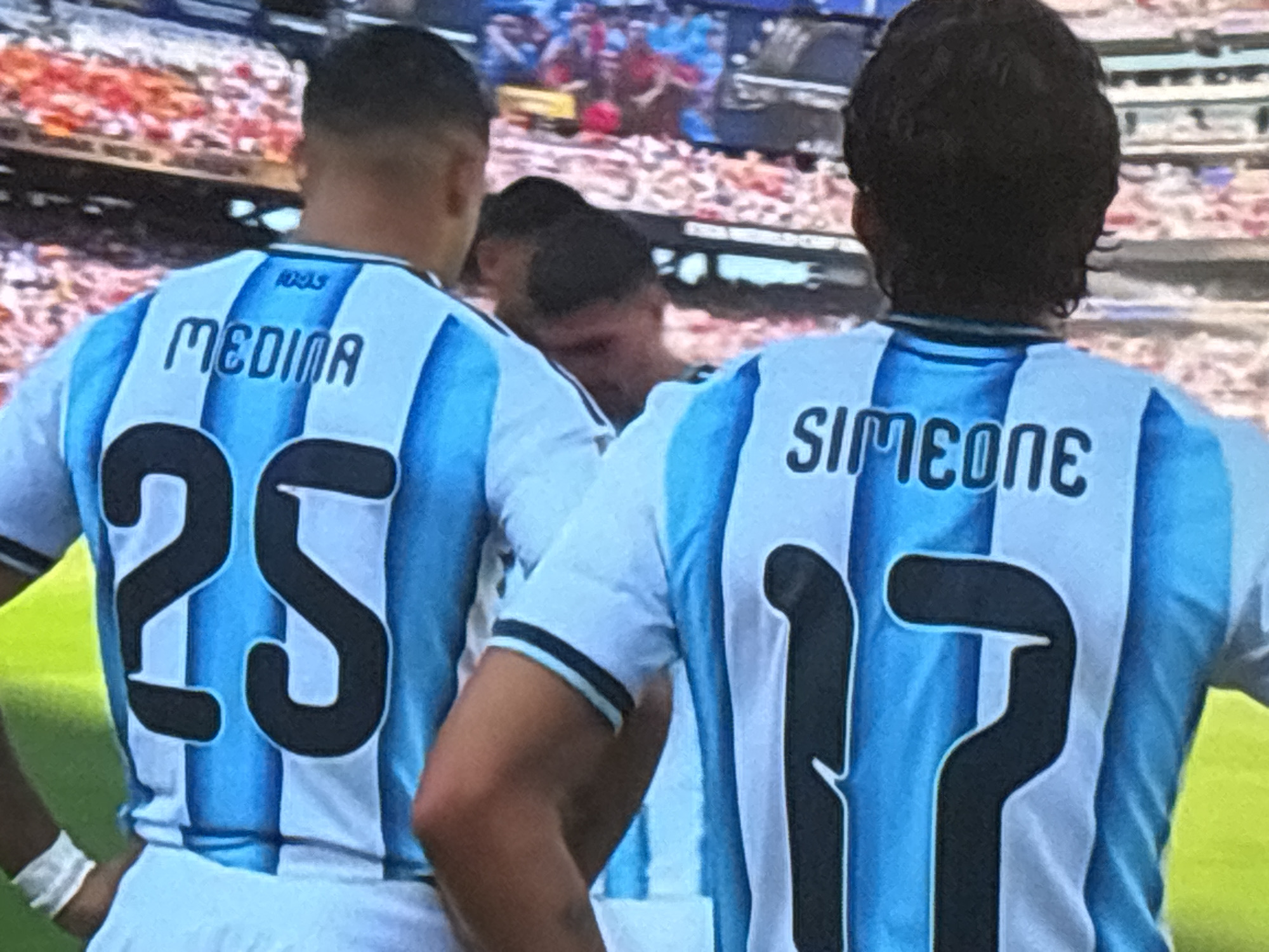 The image shows the backs of two football players wearing light blue and white striped jerseys. The player on the left has the name "MEDINA" and the number "25" on his jersey. The player on the right has the name "SIMEONE" and the number "17" on his jersey. The players are in what appears to be a stadium, with blurred spectators in the background. The lighting suggests it is daytime. The players are facing away from the camera, and their torsos and backs are visible. There is no indication in the image that this scene is located in Burgas, Bulgaria.